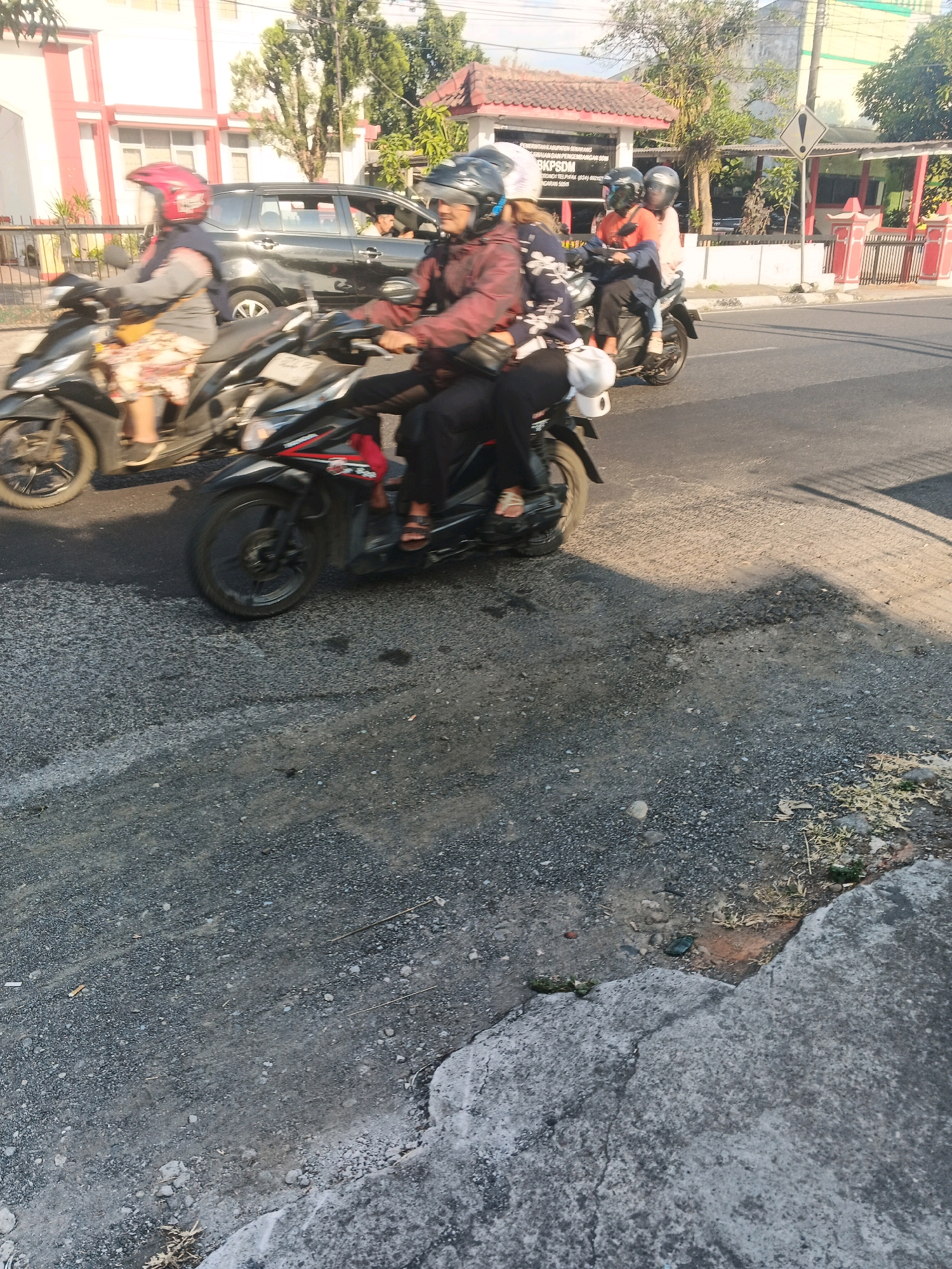 In Ungaran, Indonesia, a motorcycle with two occupants is seen in motion on a paved road. The rider wears a red helmet and a gray jacket, while the passenger has a white helmet and a patterned jacket. The motorcycle is black with red accents. A second motorcycle, also with two occupants, is visible behind and to the left of the first. The passenger on this second motorcycle wears a dark helmet and a patterned jacket, and the rider wears a light-colored helmet and a red jacket. A black car is parked to the left of the road, and a building with a white facade and red trim stands behind it. The road surface is asphalt with visible gravel and some patches of wear. The environment appears to be an urban or semi-urban setting with trees and a building in the background. The lighting suggests it is daytime, possibly late afternoon due to the shadows cast on the road.