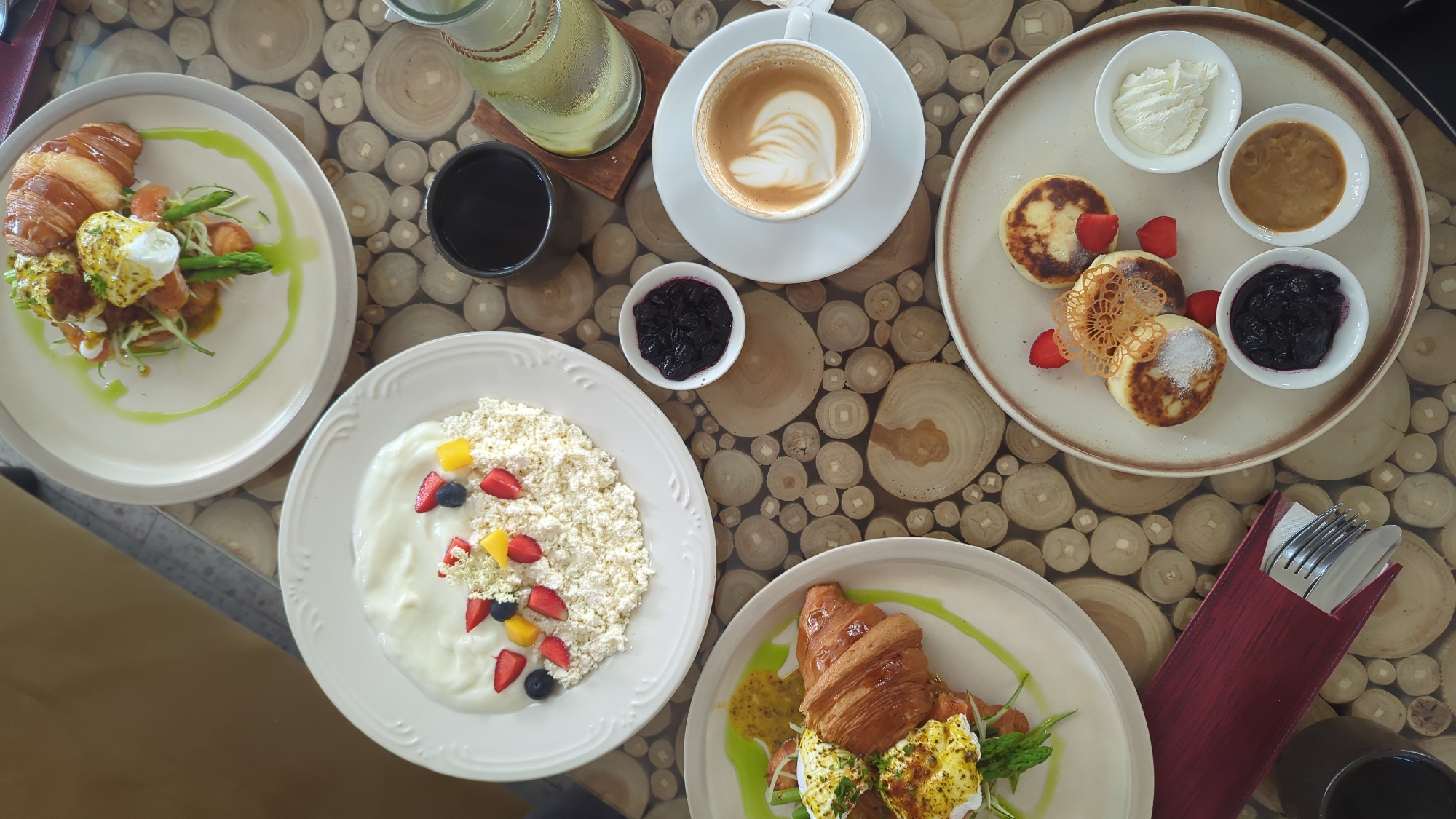 This overhead shot presents a vibrant breakfast or brunch spread laid out on a distinctive wooden table. The table's surface is composed of numerous light-toned, circular log cross-sections, creating a natural mosaic pattern.
The meal features several dishes and drinks. On the left, two similar plates each hold a golden croissant topped with poached eggs, fresh green asparagus spears, and a drizzled green sauce, resembling a gourmet eggs Benedict. Towards the bottom left, a white plate is filled with what appears to be cottage cheese or ricotta, alongside a generous swirl of yogurt. This dish is brightly garnished with diced strawberries, blueberries, mango or peach pieces, and delicate white flowers.
In the upper right, a white coffee cup with artistic latte foam sits on a saucer. Nearby, a clear glass bottle of water, infused with lemon and adorned with twine, adds a refreshing touch, alongside a small black cup, likely containing tea or coffee.
The main sweet dish, positioned in the upper right, showcases three golden-brown pancakes or syrniki, lightly dusted with powdered sugar, and decorated with fresh strawberry slices and a delicate, intricate sugar crisp. This plate is accompanied by three small white bowls: one holding creamy whipped cream or sour cream, another a rich caramel-colored sauce, and a third containing dark blueberry jam. An additional small bowl of blueberry jam is visible near the cottage cheese dish.
The dining setup, with multiple plates and drinks, suggests a meal for at least two people. The bright, natural lighting indicates it is daytime. Silverware is neatly presented in a red fabric holder in the lower right. The appealing presentation and variety of dishes suggest a pleasant dining experience in a cafe or restaurant setting, aligning with the North Kuta, Indonesia context. There is no visible text in the image.
