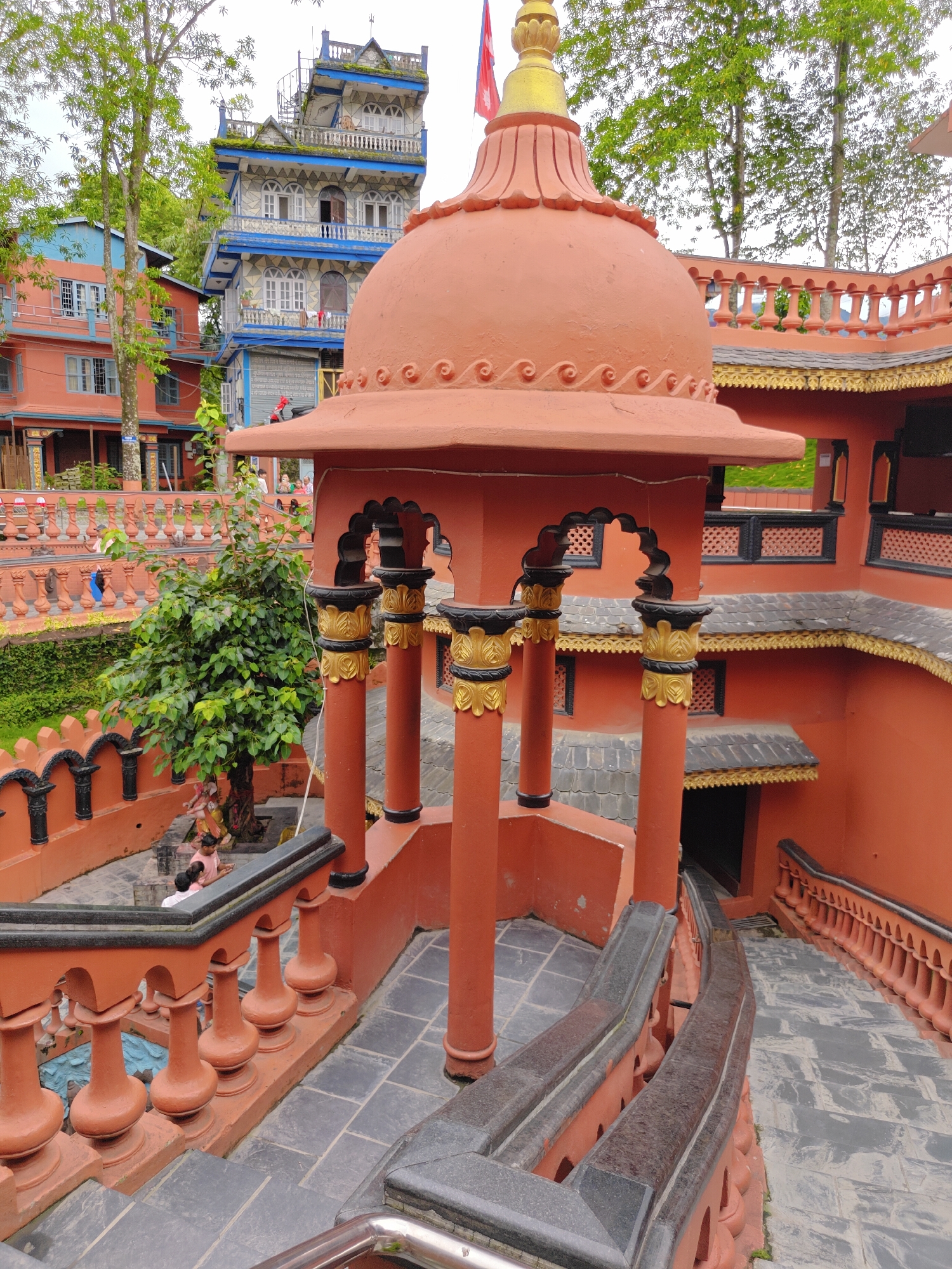 This image shows a view of the World Peace Pagoda in Pokhara, Nepal. In the foreground, an orange stupa with a gold finial stands prominently. Red-orange walls and ornate railings with black and gold accents frame a descending stone staircase. A small tree with green foliage grows within a courtyard area visible in the middle ground. In the background, a tall, multi-story building with blue and white detailing and a spiral staircase on its exterior is visible, along with other colorful buildings and lush green trees. A red flag flies from the top of the multi-story building. Several people are present in the scene, including individuals walking on the stairs and in the courtyard. The sky is overcast.
