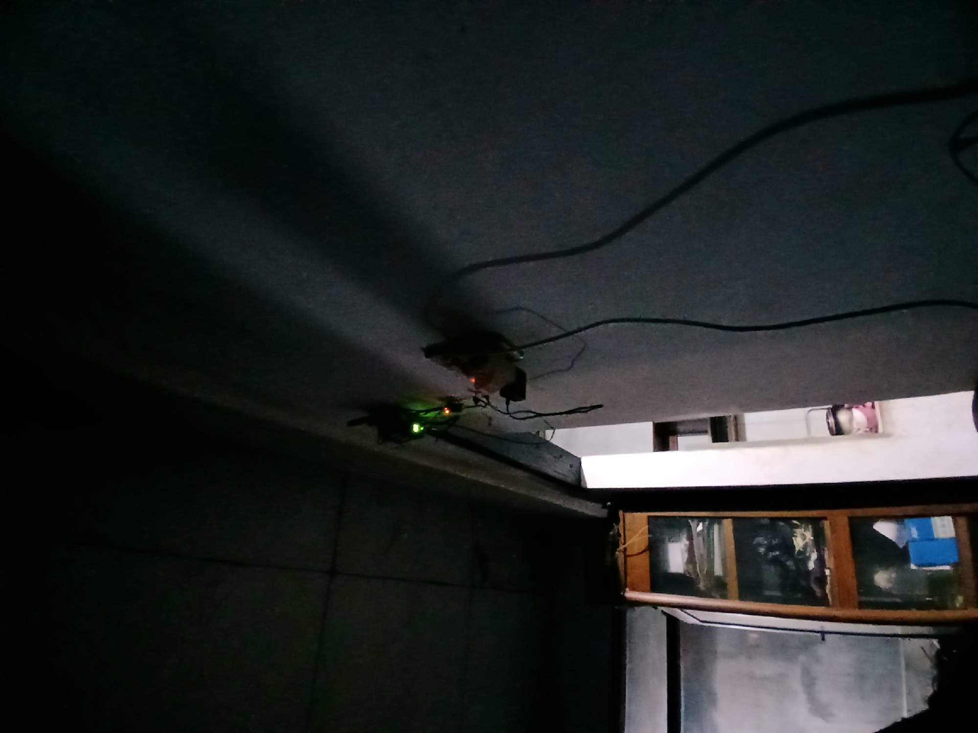The image depicts an indoor scene, likely a room, with predominantly dark tones. In the upper left portion, the ceiling is visible, exhibiting lines that suggest panels or structural elements. On the right side of the image, a wall extends upwards, with a noticeable shadow cast across it from an unseen light source. Attached to the wall, near the ceiling and overhanging edge, is a power strip with several plugs, illuminated by small green indicator lights. Wires are seen extending from the power strip, trailing downwards and to the right. In the lower left, a tall, narrow wooden cabinet with glass doors is visible. The cabinet contains several shelves with various items, including what appear to be books or files on the upper shelves, and boxes or containers on lower shelves. The background behind the cabinet is obscured by darkness, but some light appears to be emanating from a possibly open doorway or window on the far left. The overall impression is that of a dimly lit interior space containing electrical devices and storage furniture.
