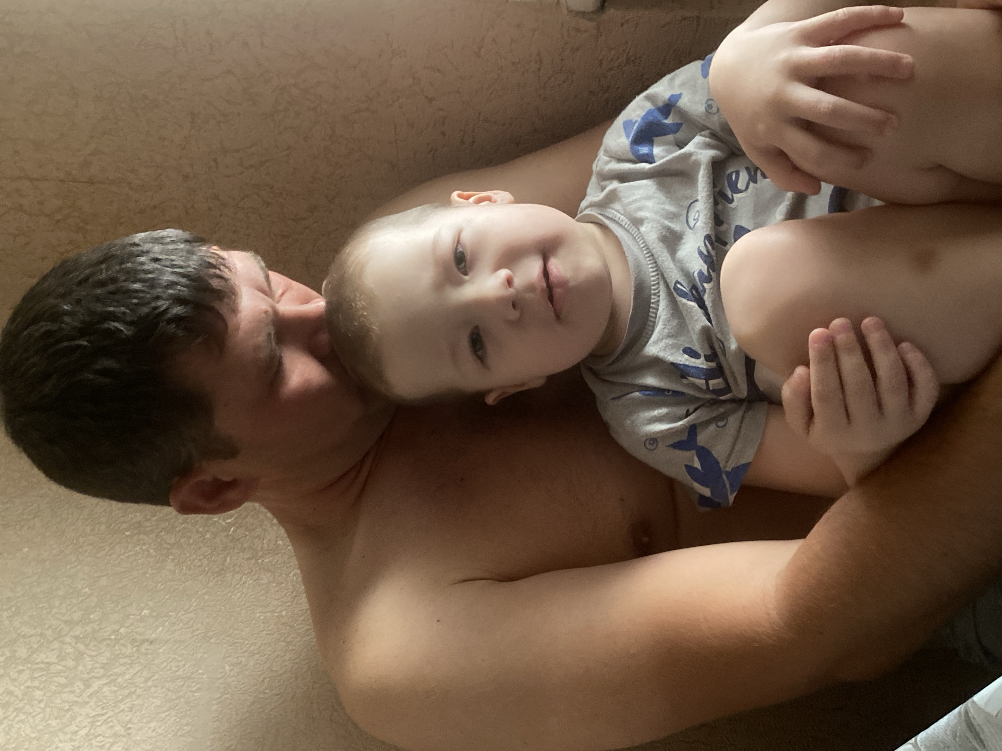 A man lies shirtless, holding a young child wearing a gray shirt with blue sharks. The man's dark hair is closely cropped, and his skin tone is light. The child's face is turned towards the camera, showing a gentle smile. The background is a textured, neutral wall. The image does not contain sufficient information to determine the location in Sochi, Russia.
