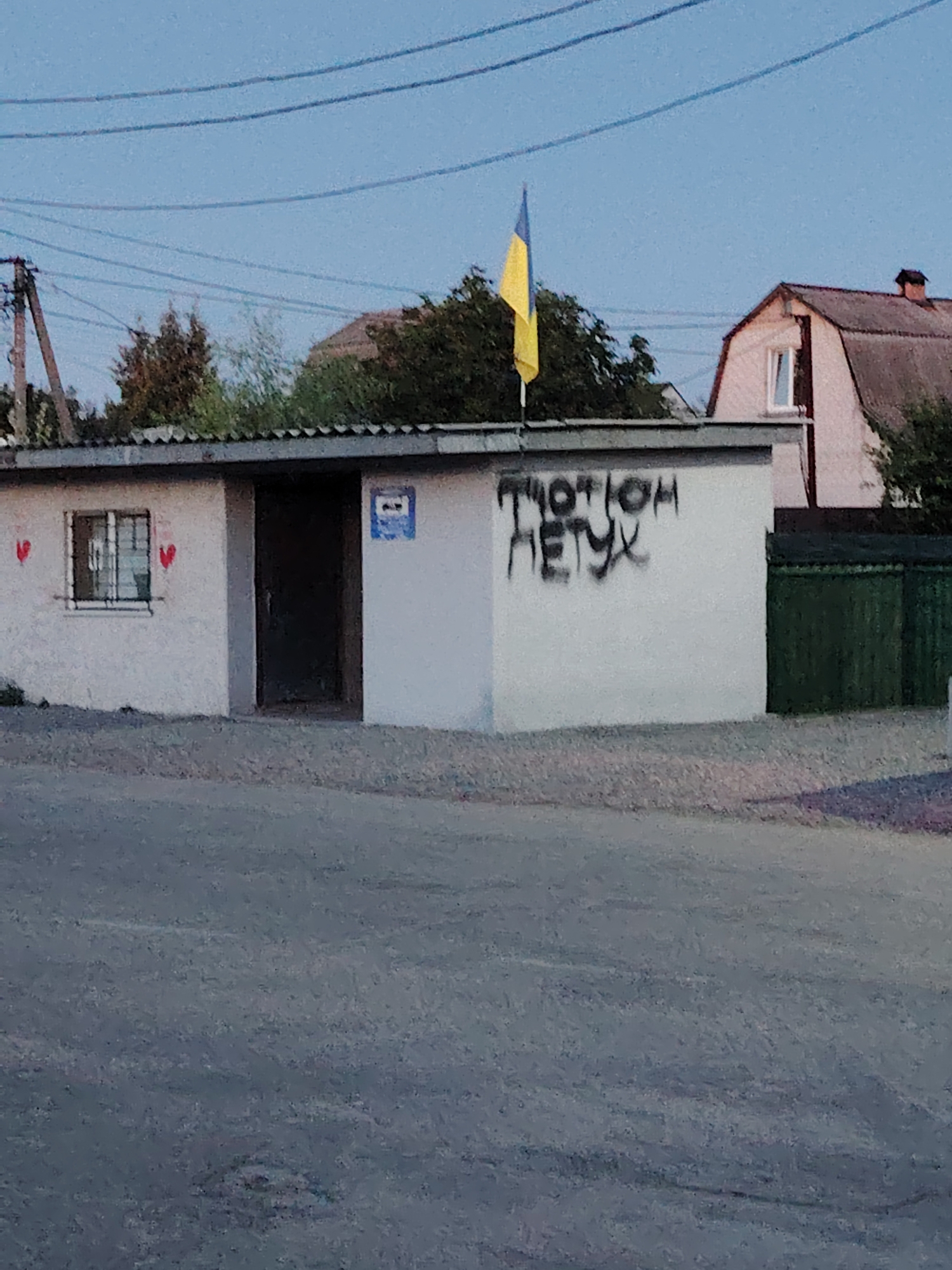 The image shows a small, white, rectangular building with a dark door and a window. The building is located in the city of Oseshchyna, Ukraine. There is graffiti on the right side of the building that reads "TUOTIOM HETYX". A Ukrainian flag is flying above the building. In the background, there are trees and houses. The sky is clear and blue. The image is taken from a low angle, looking up at the building.