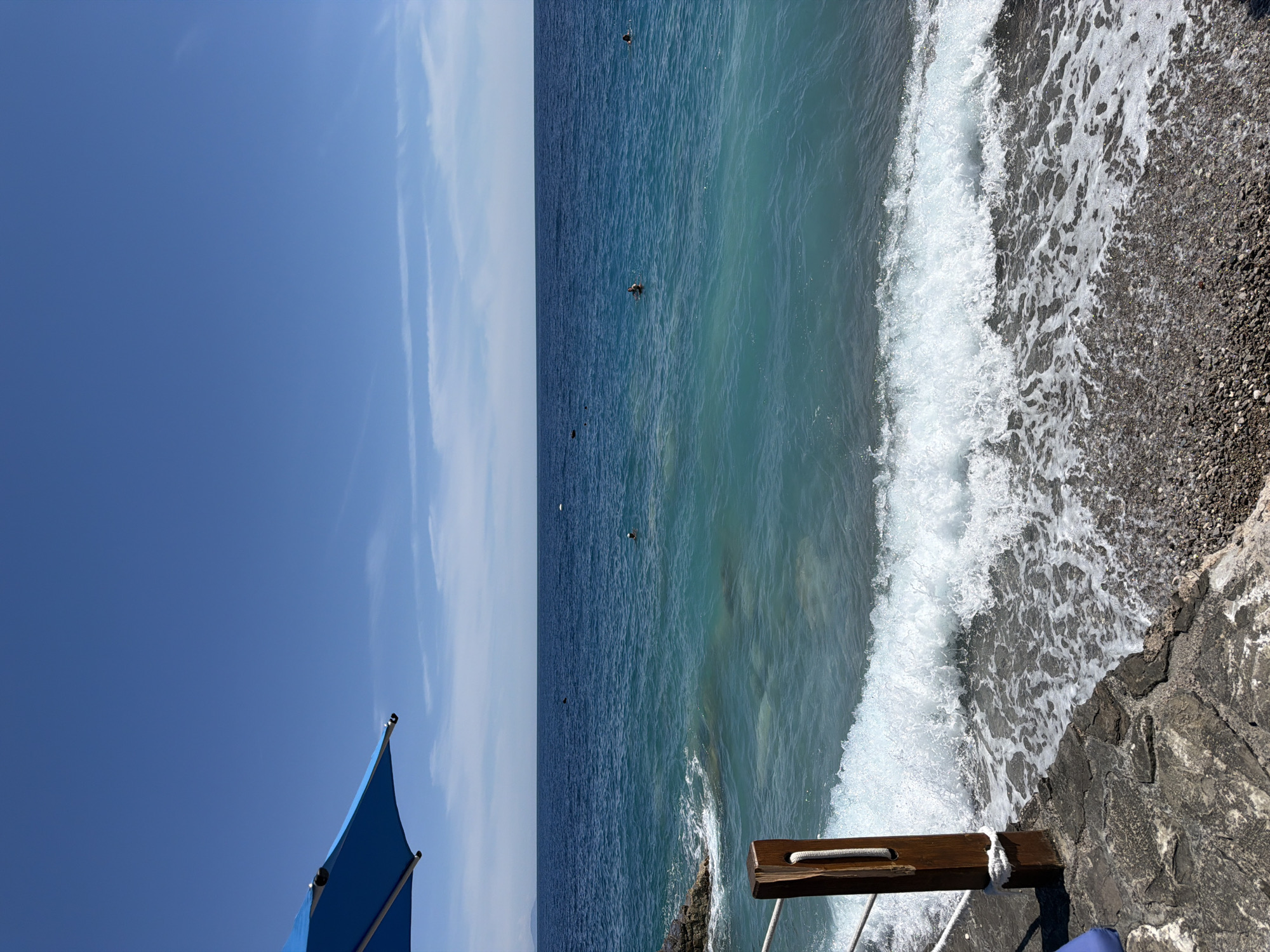 A vibrant blue umbrella is partially visible in the top left corner of the frame. Below it, a rope barrier separates a rocky, pebble-strewn shoreline from the tranquil, turquoise waters of the sea. The ocean stretches to the horizon, meeting a clear, bright blue sky dotted with thin, wispy clouds. Several small, dark figures can be seen swimming or wading in the distance, their presence indicating a leisurely day by the water. The waves gently lap at the shore, creating a soothing rhythm and a sense of peaceful seclusion. The scene evokes a sense of a secluded beach cove within the picturesque setting of Prijevor, Montenegro.