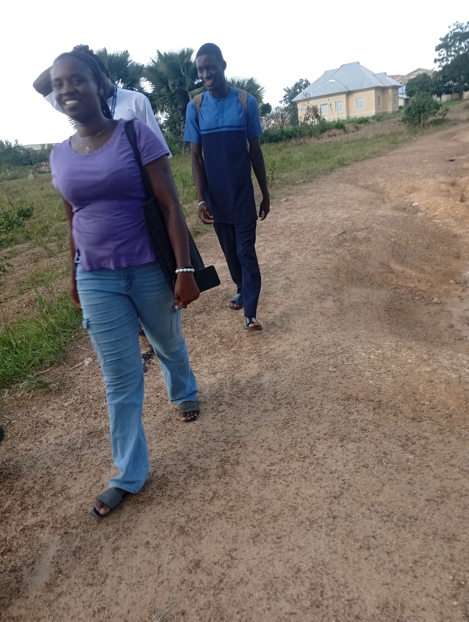 Two individuals are walking on a dirt path. A woman in the foreground is wearing a purple t-shirt and blue jeans, and a black bag is slung over her shoulder. She is smiling and looking towards the camera. Behind her, a man in a blue and dark blue t-shirt and dark pants is walking away from the camera. He is wearing a backpack. In the background, there is a light yellow house with a grey roof, surrounded by greenery and palm trees. The scene appears to be in an outdoor, possibly rural or suburban, setting in Rafin Par, Nigeria.