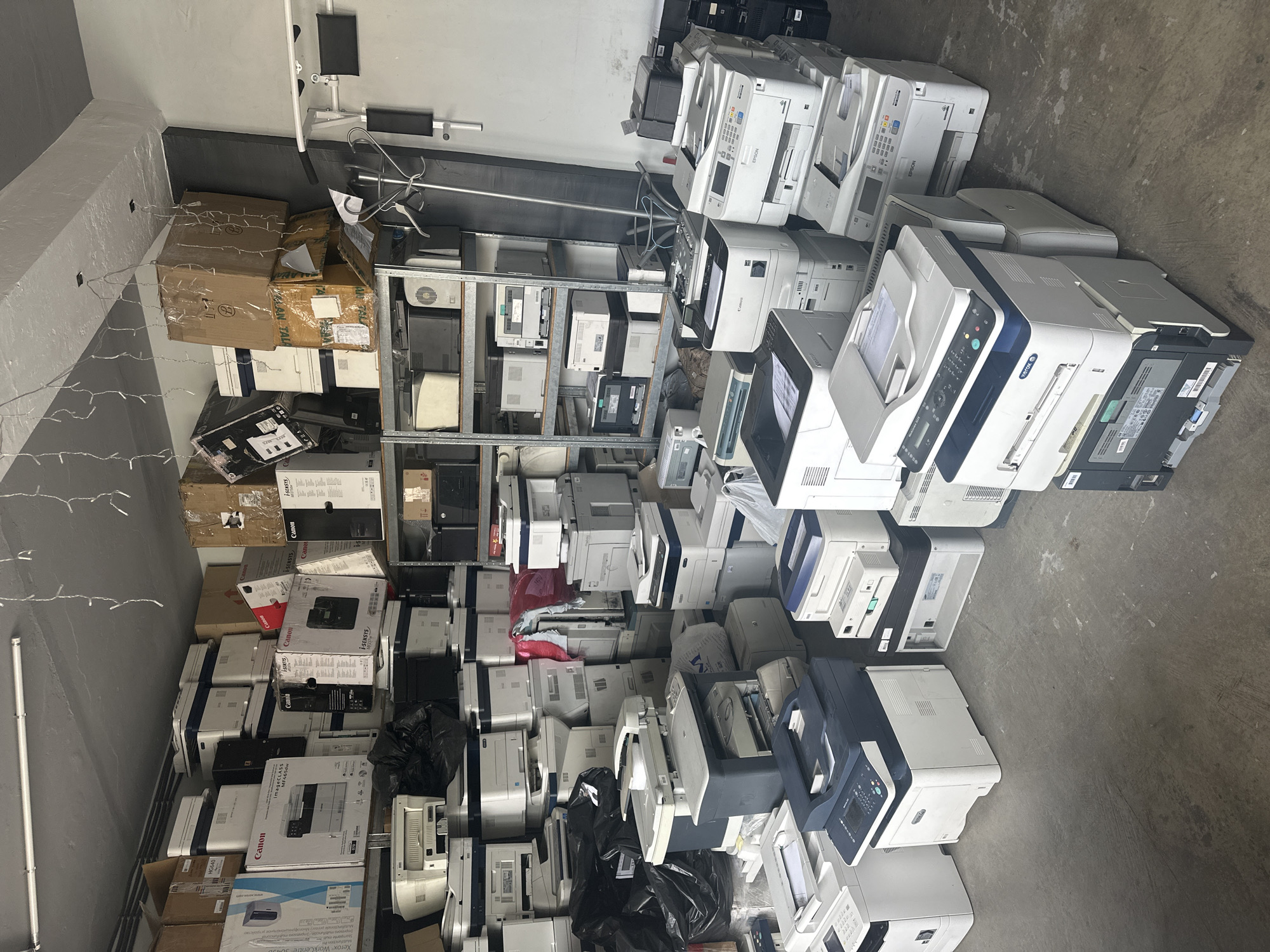 The image shows a large, cluttered storage space in Unknown, Unknown, likely a warehouse or commercial building. The room has gray walls and a gray concrete floor. The dominant feature is the dense accumulation of numerous printers and photocopiers. The devices vary in model and condition, from seemingly new in boxes to used or disassembled. Some are stacked precariously on shelves, while others are scattered directly on the floor. Some printers are still in their original packaging from Canon and Xerox. There are also several black plastic bags interspersed among the electronics, possibly containing waste materials. A metal shelving unit stands along one wall, filled with smaller electronics and components, as well as some cardboard boxes. A strip of light is present along the top of the wall, adorned with string lights. The overall impression is that of a recycling or storage facility dealing with a surplus of electronic waste.