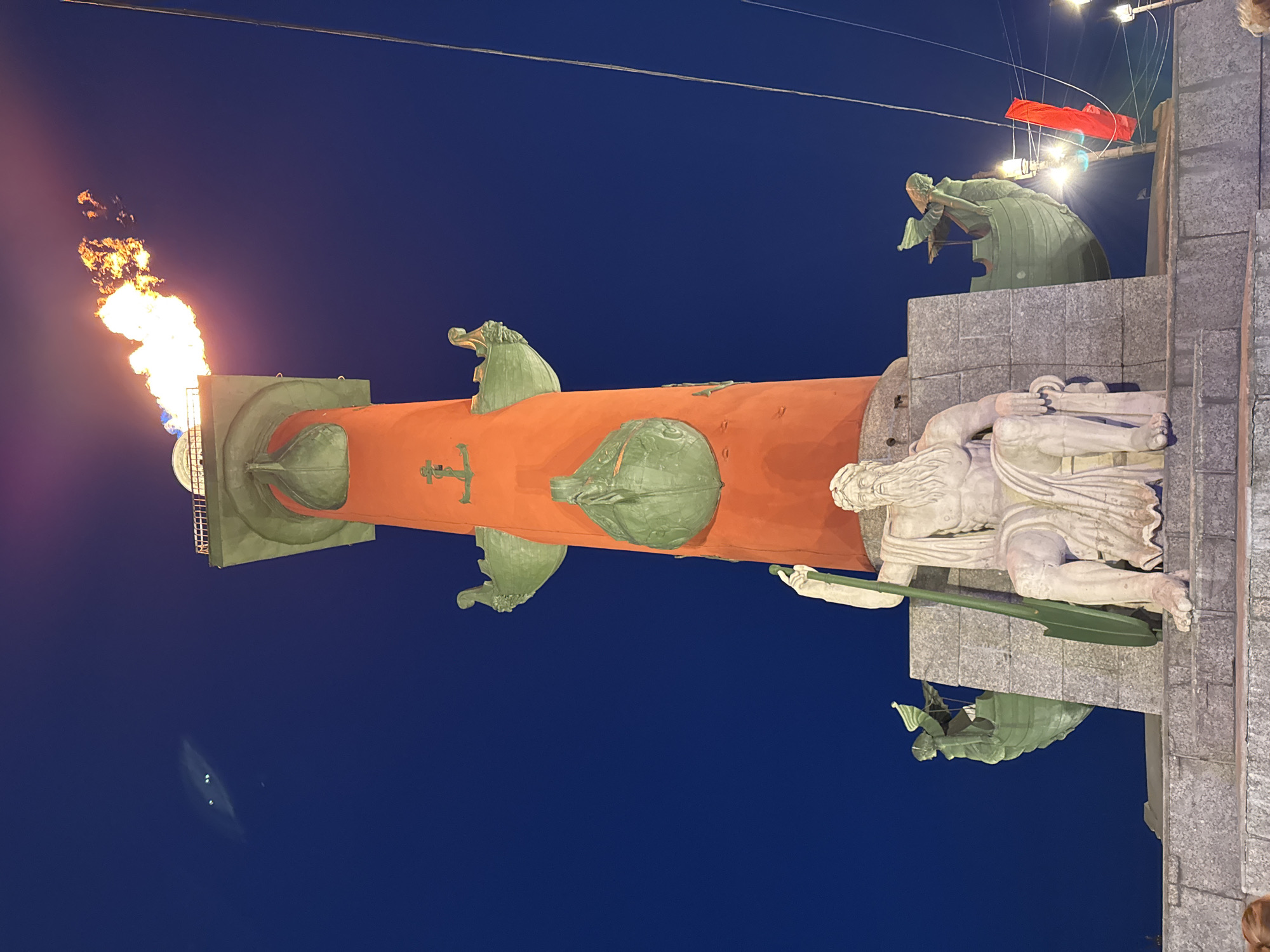 The image captures the Rostral Column, a significant landmark in Saint Petersburg, Russia, against a deep blue evening sky. At the top of the column, a large orange flame is ignited, emitting a bright glow. The column itself is decorated with green-colored naval prows and a small green cross. At the base of the column, sculptures and carved stonework are visible. Additional decorations and structures are situated to the right of the frame and are adorned with several white lights and a red cloth.