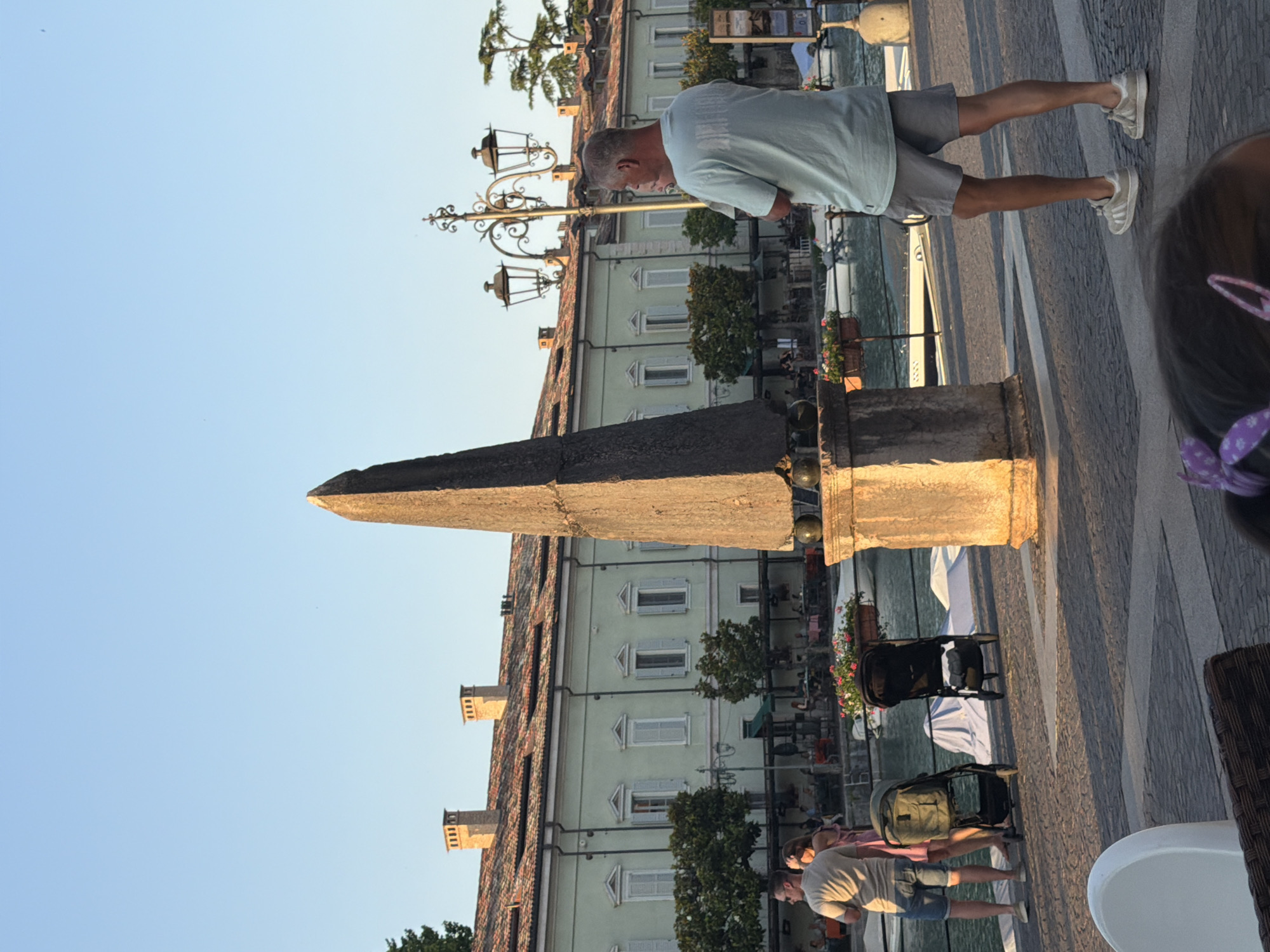 The image depicts a sunny day in Peschiera del Garda, Italy, likely in the late afternoon or early evening given the warm, long shadows cast by the structures. The scene is set along a waterfront promenade with a cobblestone walkway featuring a decorative pattern of light and dark grey paving stones.
In the foreground, a man in a light blue t-shirt and grey shorts is walking away from the camera. He is wearing white sneakers. To the right of the man, a young child's head is visible, with a purple hair accessory. Further down the promenade, another couple is walking, the woman in a pink dress and the man in a light-colored t-shirt and blue shorts. A baby stroller is visible behind them.
Dominating the mid-ground is a light green building with a terracotta tiled roof. The building has multiple windows with light blue shutters, and several chimneys. In front of the building, a wrought-iron lamppost with two ornate lamps stands tall. A dark, obelisk-shaped monument or sculpture is also present in the mid-ground, casting a shadow.
The background shows a canal or body of water with several boats docked. Across the water, more buildings can be seen, some with balconies and potted plants. The sky is clear and blue, indicating good weather. The overall atmosphere seems relaxed and leisurely, suggesting a typical day of vacation or sightseeing in a European town. No visible text is present in the image.