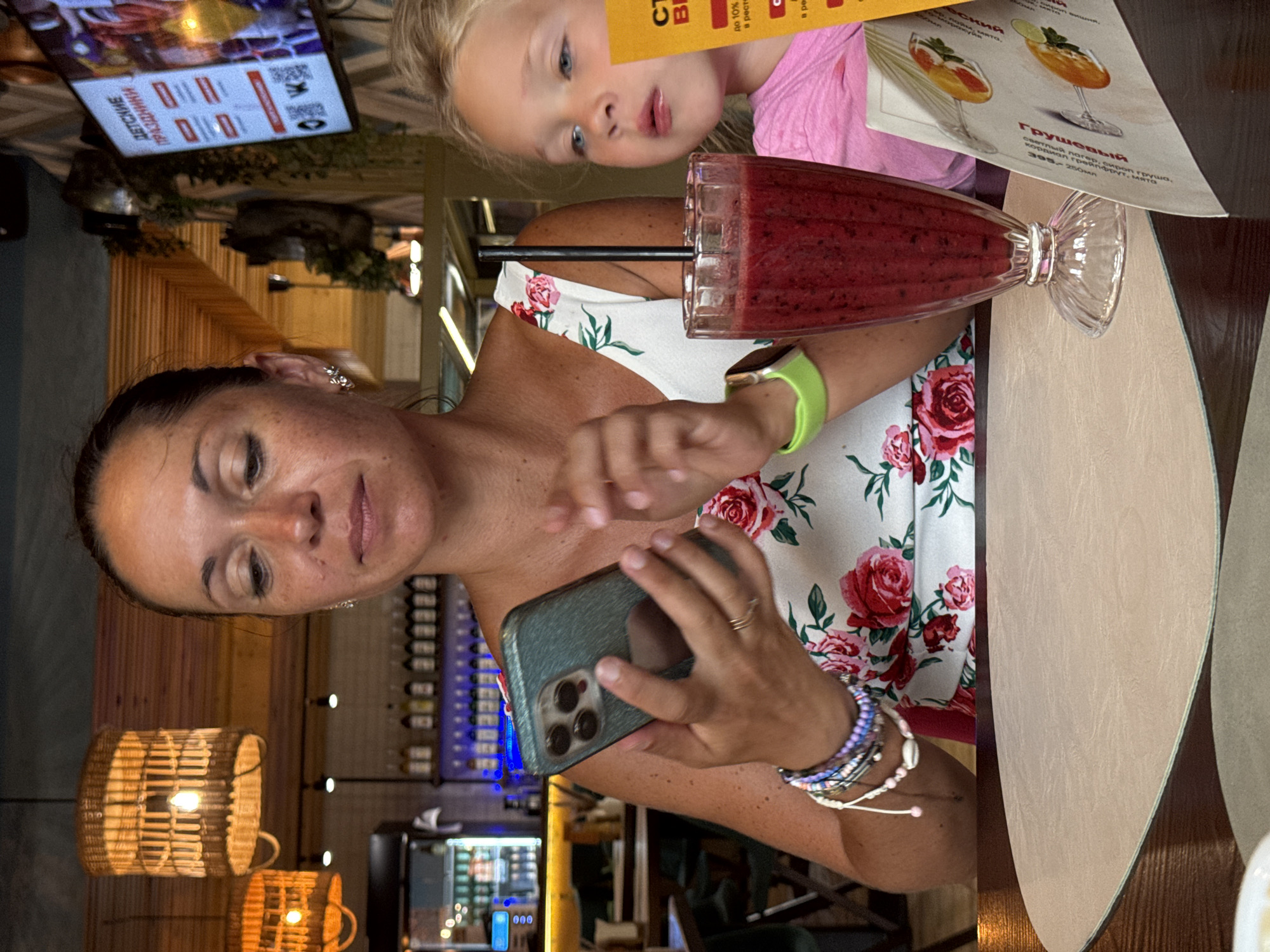 In Sochi, Russia, a woman is sitting at a table in a dimly lit restaurant, holding a smartphone. She wears a white dress with a floral pattern and several bracelets on her left arm. To her right, a young girl with blonde hair is looking to the side. A glass of red smoothie with a straw is on the table in front of them, next to a menu. The background shows wooden panels on the ceiling and walls, with lamps and bottles of alcohol on shelves. A large screen displaying text in Russian is also visible on the wall.