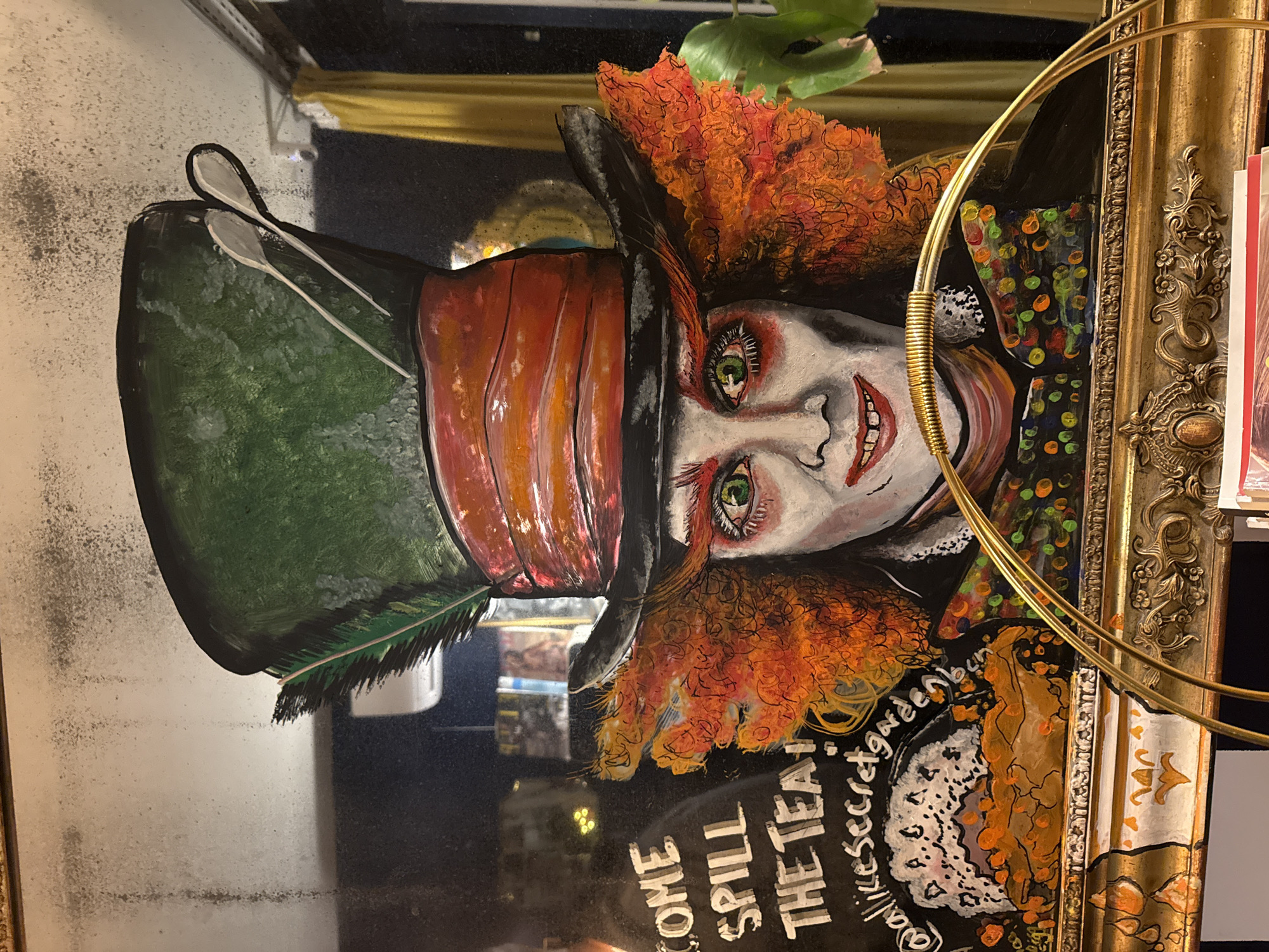 A framed mirror reflects an artistic representation of the Mad Hatter from Alice in Wonderland. The character is depicted with a large green hat, topped with spoons, and a red striped band, set at an angle. His orange hair frames a pale face with green eyes and a sinister smile. A black patterned jacket completes the character. Text on the mirror reads, "COME SPILL THE TEA!" and "@alikesecretgardenbcn," suggesting the artist is named "Alike Secret Garden," and "bcn" is the commonly used abbreviation for Barcelona. The frame itself is ornate and gilded. The image's surroundings are not clearly visible due to the reflection, though a gold wire object is partially visible, possibly the mirror's frame.