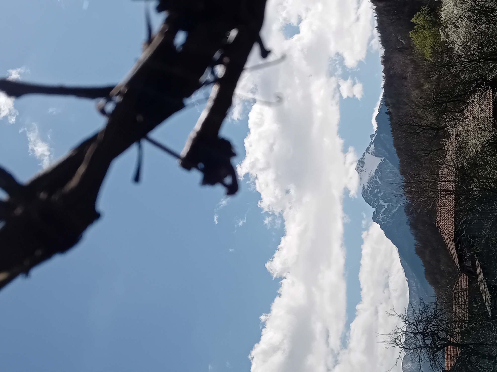 The image shows a view of a mountain with snow on its peak, partially obscured by clouds in a bright blue sky. In the foreground, there's a blurry, dark, skeletal structure of what appears to be a wooden bridge or scaffolding, possibly in disrepair. The right side of the image shows a treeline and buildings with red tile roofs, suggesting a rural or mountainous area. The photograph is taken from a low angle, looking upwards. Location: Unknown, Unknown.