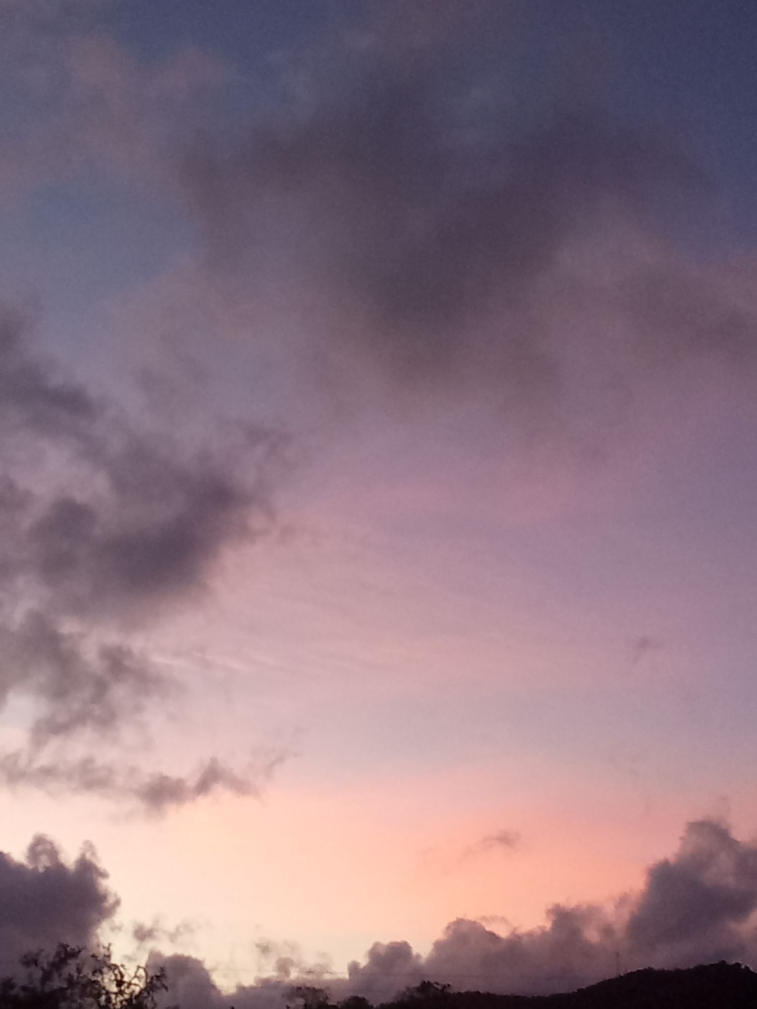 The image displays a twilight sky observed from Juan Griego, Venezuela. The sky exhibits a prominent color gradient, transitioning from a dark blue-grey at the top, through shades of purple and lavender in the mid-section, to a distinct peach-orange hue along the lower horizon. Scattered, irregular cloud formations are present, appearing dark grey and dense in the upper left, mid-left, and lower right areas, while lighter, wispy clouds are visible against the colorful sky backdrop. Along the very bottom edge of the frame, a dark, indistinct silhouette of what appears to be distant vegetation, likely trees, and possibly a low-lying hill or landmass is visible against the lighter sky. There are no discernible human figures, man-made structures, or specific objects, nor any apparent actions or interactions within the scene.