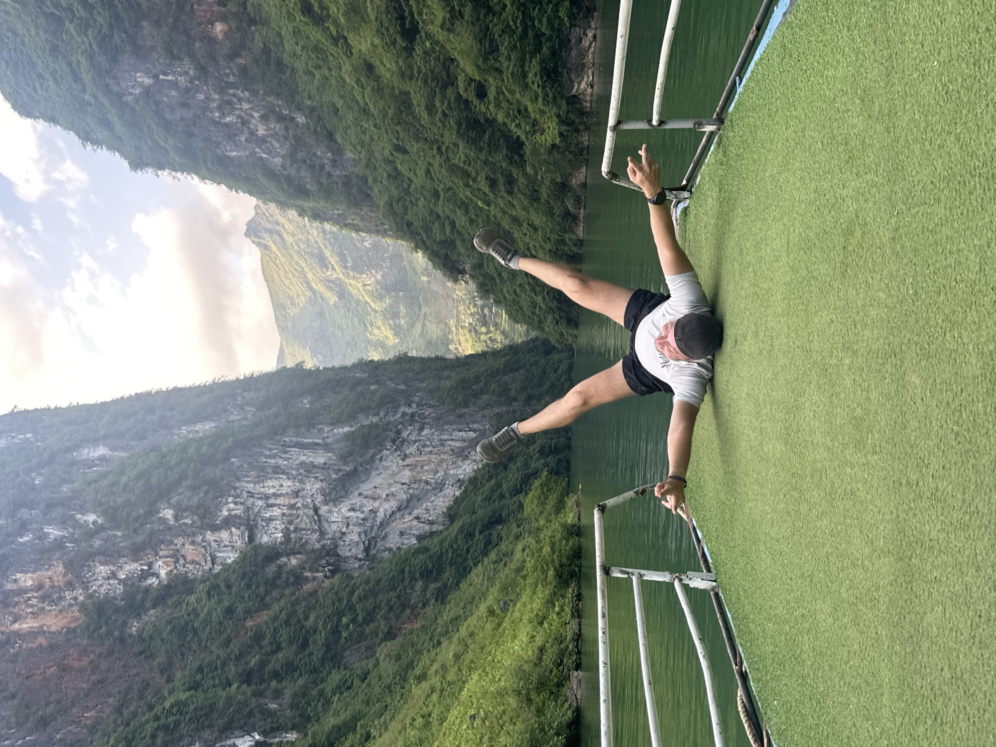 A man is suspended horizontally on a boat against a backdrop of mountains, which places the location in the city of Đồng Văn Commune, Vietnam. He grips the boat's railing with both hands, his body parallel to the deck. He wears a gray t-shirt, black shorts, and gray athletic shoes. His legs are spread in a "V" shape. The deck is covered in artificial green turf. The boat is sailing on a body of water. In the background, steep, tree-covered mountains rise on either side, with a cloudy sky visible above.