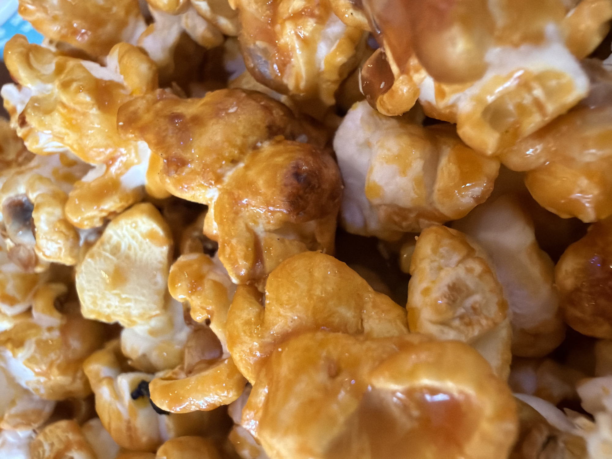 A close-up shot reveals a collection of caramel popcorn pieces. The popcorn is coated in a shiny, golden-brown caramel, with some pieces having a darker, toasted appearance. The texture is visibly irregular, with crevices and protrusions coated in caramel. The image suggests an indoor setting and a focus on the caramel popcorn. The location is not discernible from the image.