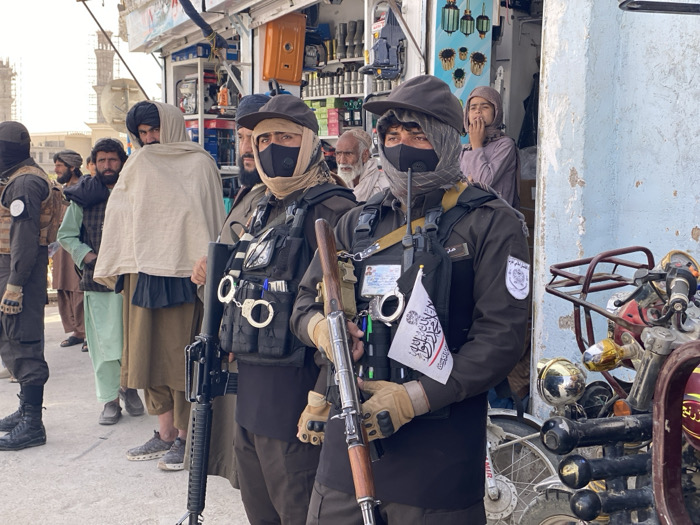 The image depicts a street scene in Kandahar, Afghanistan. In the immediate foreground, two armed men in dark uniforms and tactical vests stand prominently. The man on the right wears a black face mask, a light-colored head covering under a dark cap, and brown gloves. He holds a rifle with a wooden stock in his gloved hands and has a small white flag bearing black Arabic script (the Shahada) attached to his tactical vest, which also displays handcuffs and an identification badge. The man to his left, slightly behind, wears similar attire including a black face mask, head covering, and dark cap, and carries a dark rifle. His tactical vest also features handcuffs.
To the left of these armed individuals, a group of civilian men in traditional Afghan attire, including turbans and shawls, stand and observe. One man wearing a dark turban looks directly towards the viewer. Another man is draped in a white shawl. Further left, partially obscured, is another individual in a dark outfit with a similar head covering.
On the far right, multiple motorcycles are parked against a light-colored, dilapidated wall with peeling paint. Behind the armed men, a shop facade is visible, displaying various merchandise such as tools, light fixtures, and household items. A young man in a light-colored tunic stands in the shop's doorway, looking out. In the distant background on the far left, two distinctive minarets from a large architectural complex are visible against the sky. The ground is paved and light-colored.