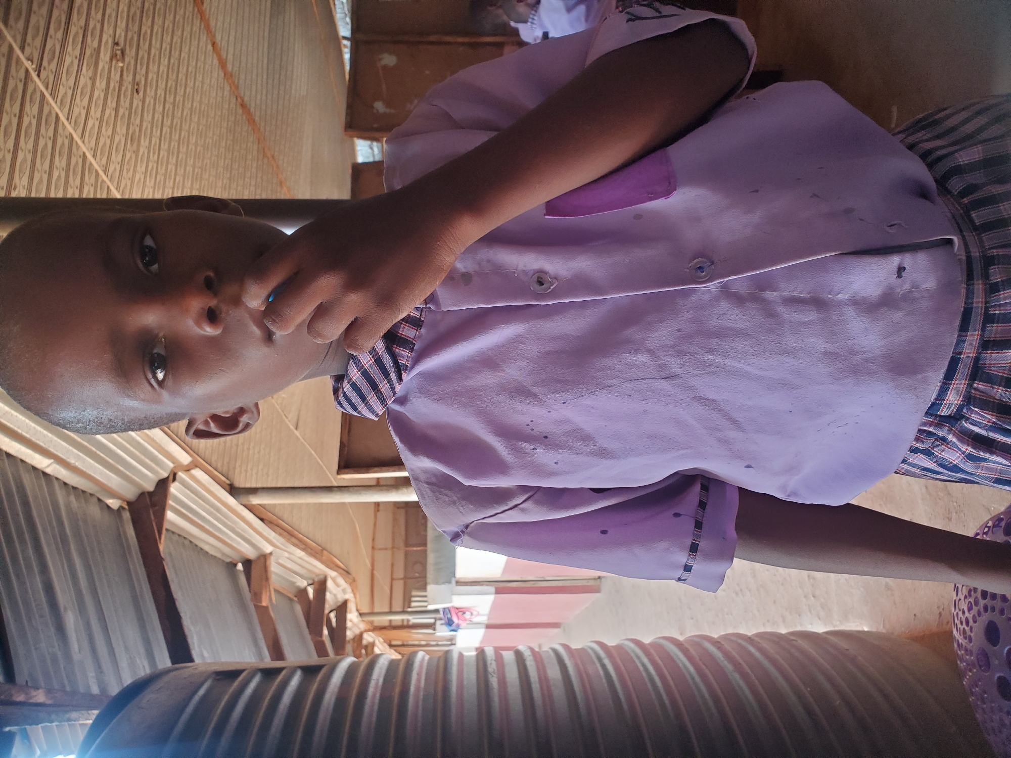 A young child, likely a boy, with dark skin and short hair, is the central subject, looking directly into the camera with a neutral expression. He is wearing a light purple collared shirt with a darker purple patch/pocket, and a plaid uniform bottom in red, blue, and white checks. The shirt exhibits several dark stains or marks. One of his hands is raised near his mouth or chin.
The scene appears to be an outdoor or semi-enclosed setting, consistent with a school environment or a covered area in Ukwu-Abwa, Nigeria. The background reveals a structure with corrugated metal roofing and wooden beams, suggesting simple construction. In the foreground, a large, dark, ridged cylindrical object, possibly a water pipe or storage tank, spans the bottom of the frame. To the child's right, a purple, perforated object, resembling a plastic basket or stool, is partially visible. Bright, natural daylight illuminates the scene, indicating it is daytime. A blurred figure, possibly another child, is vaguely discernible in the far background. There is no visible text in the image.