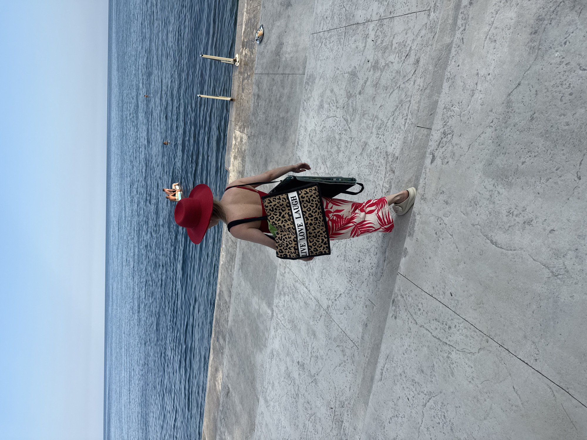 A person wearing a red hat, red bathing suit, and a white and red leaf-patterned skirt walks down concrete steps toward the sea. The person is carrying a leopard-print tote bag with the words "LIVE LOVE LAUGH" printed on it in white lettering, and a green bag. The sea is calm and blue, with a few people visible in the water and on an inflatable raft. Two white poles stand in the water near the shore. The sky is clear and blue. The location is identified as Utios, Ukraine.