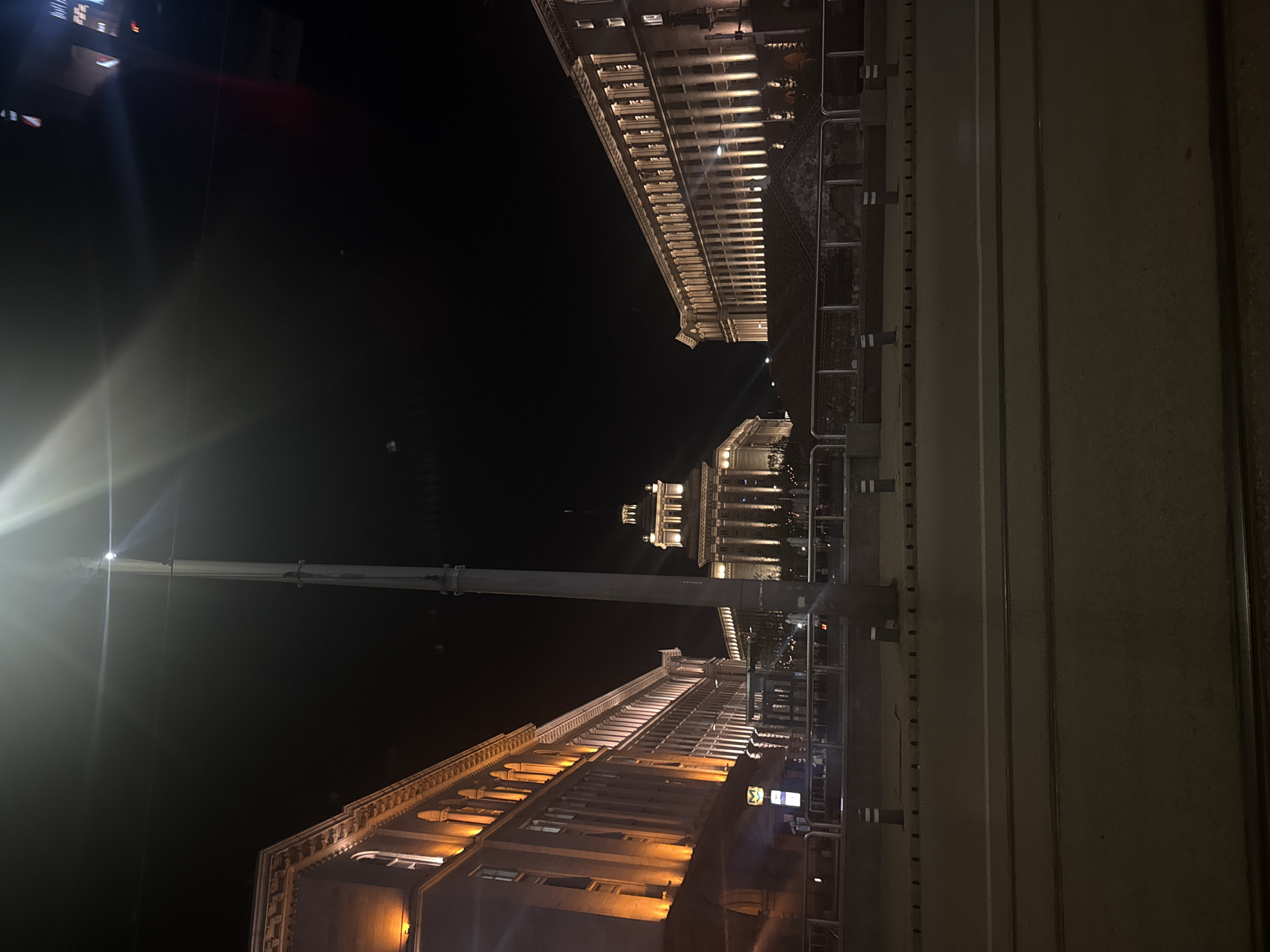 This nighttime image captures a grand urban scene in Sofia, Bulgaria, characterized by imposing classical architecture under dramatic artificial lighting. The scene unfolds across a wide street or square, flanked by stately buildings.
On the left side, a large, multi-story building with ornate cornices and columns is illuminated from below with warm, golden lights, highlighting its intricate facade. On the right, another similarly styled building, also well-lit, mirrors the architectural elegance. In the background, at the far end of the square, stands a majestic building, possibly the Council of Ministers or part of Sofia's Largo complex. This structure features a prominent, columned portico and a central tower, all brilliantly lit against the dark sky.
A very tall, slender street light pole rises vertically on the left side of the frame, its powerful light source creating a significant lens flare. The paved area in the foreground and stretching into the distance shows faint lines, possibly indicating tram tracks, and is bordered by metal barriers or railings. A few indistinct figures of people can be seen near the distant buildings, suggesting pedestrian activity.
The time of day is clearly night, with the sky appearing completely black. The strong artificial lighting on the buildings and the street light create a high contrast, lending a sense of grandeur and historical significance to the location. A small sign with a white sigma (Σ) symbol on a blue background is faintly visible on one of the foreground buildings.