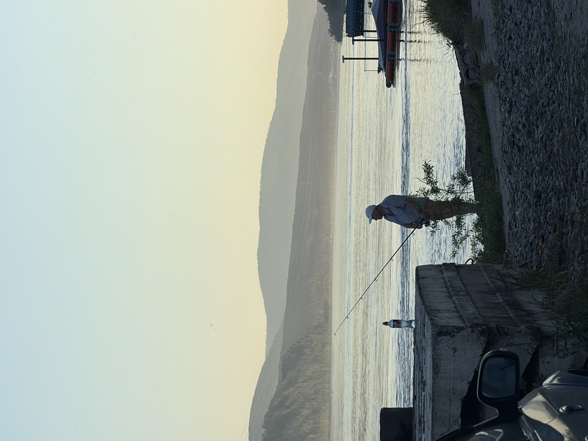 This image captures a wide riverine landscape at what appears to be early morning or late afternoon, characterized by soft, low-angle light. The scene is set along a bank of the Yenisei River near Krasnoyarsk, Russia, with mountains in the background.
In the mid-ground, a single male figure stands on the riverbank, facing right, engaged in fishing. He wears a light-colored baseball cap, a striped, collared shirt, and trousers with a camouflage or patterned design. He holds a fishing rod, the line extending into the water.
To the immediate left of the fisherman, on a large, weathered concrete block, a transparent plastic bottle with a red label is positioned. These concrete blocks, forming a substantial barrier or structure, dominate the left foreground. Partially visible in the bottom left corner is the front section of a dark-colored automobile, including a side mirror, parked adjacent to the concrete. The ground in the foreground is covered with dark, rounded pebbles and sparse green vegetation.
The river, wide and calm with gentle ripples, stretches across the middle of the frame, reflecting the pale hues of the sky. On the distant right bank, a rudimentary dock structure with two small, orange-accented pontoons or boats is visible.
The background features multiple receding layers of mountains or large hills, covered with trees, appearing increasingly hazy and desaturated towards the horizon due to atmospheric perspective. The sky transitions from a pale blue at the top to a soft yellow-orange closer to the horizon.