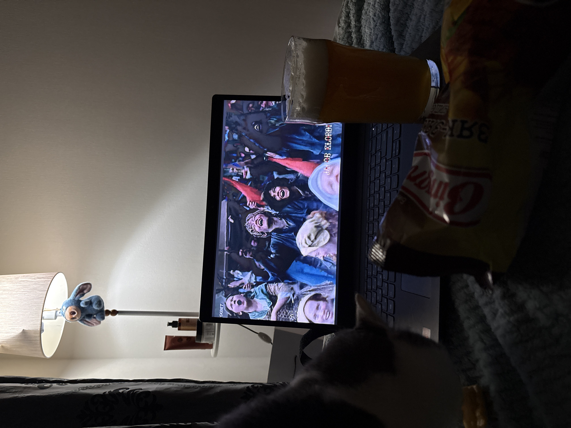 The image depicts a dimly lit indoor setting, possibly a bedroom, with a laptop screen displaying a scene from the movie "Jacob Elordi," showing a crowd of people in historical attire. A glass of beer sits precariously on the laptop, while a bag of chips, possibly "Bimbo" brand, is positioned nearby. A lamp with a decorative blue stuffed animal stands on a small shelf, along with a few toiletries. A blanket and a partial view of a cat can be seen in the foreground. There is no information to suggest that the location is Ust-Ilimsk, Russia.