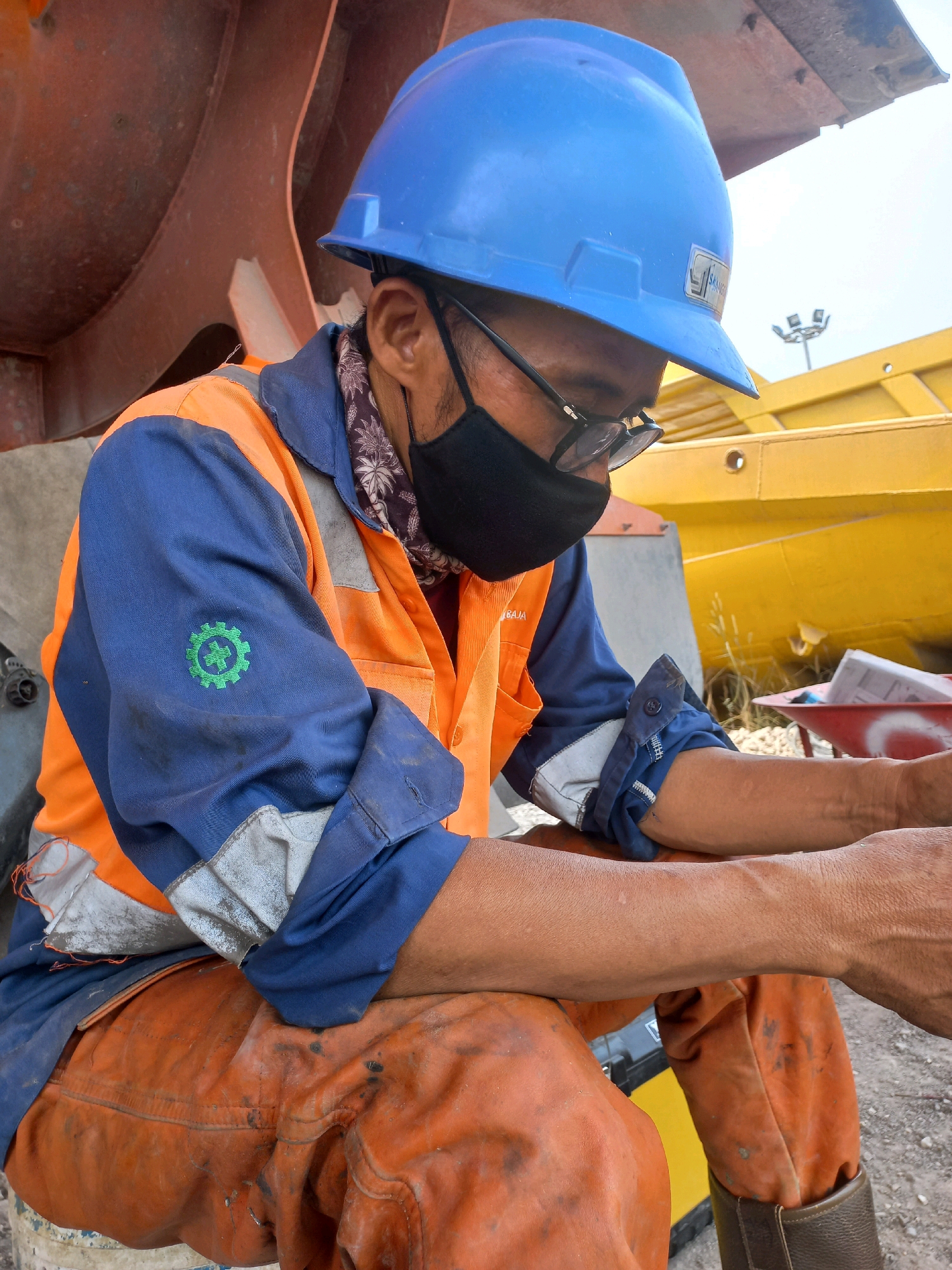 An Indonesian man wearing a blue hard hat, black face mask, glasses, a blue and orange work jacket with a green gear emblem, and orange work pants, sits outdoors. He is wearing a patterned bandana under his mask. The man's arms are resting on his legs, and his head is bowed. The background includes large, rusty metal structures, yellow industrial equipment, and a clear sky with a light fixture in the distance. The man appears to be in a work environment, possibly a construction site or industrial facility. The location is identified as Paringin, Indonesia.
