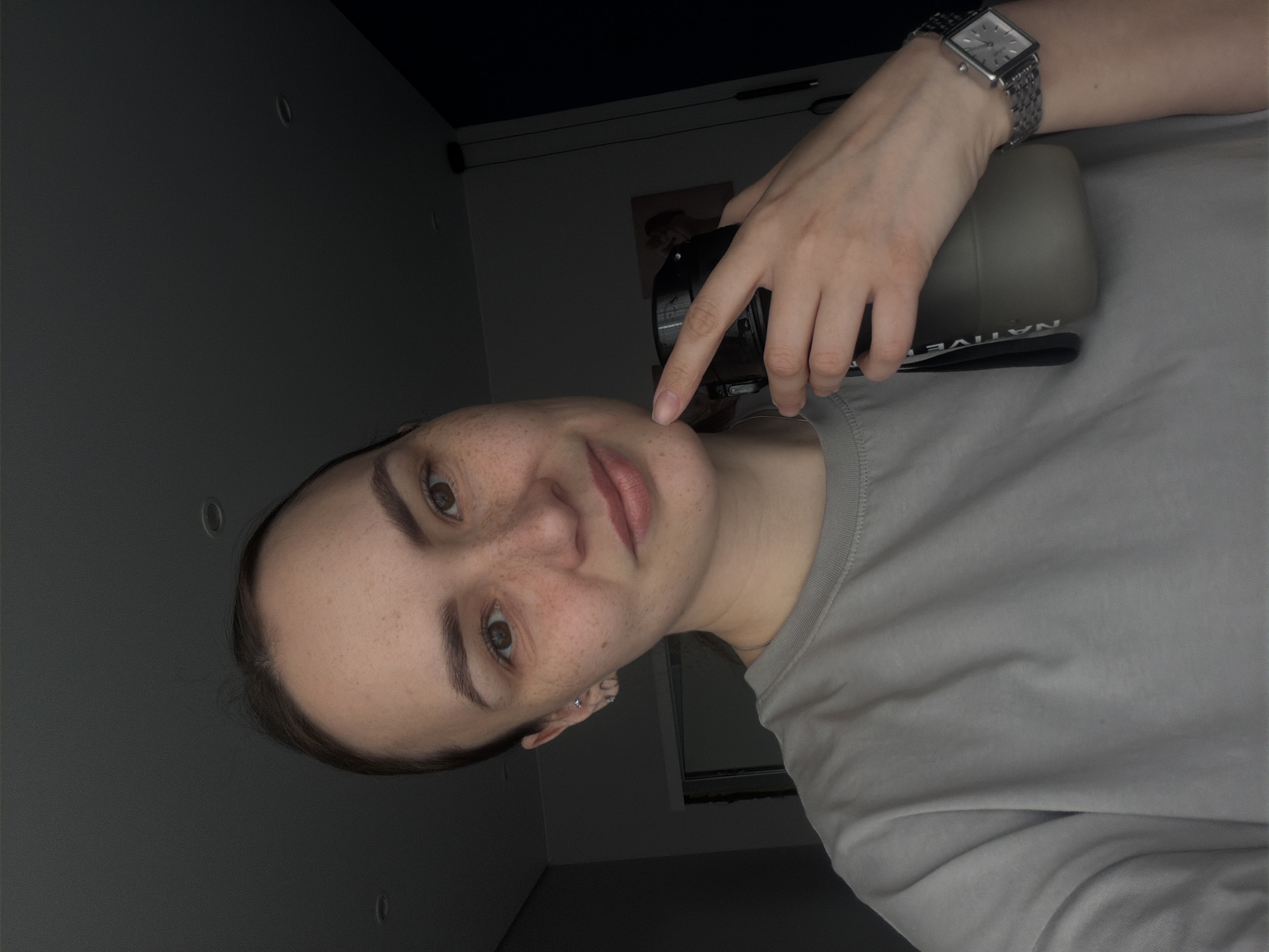 A person with freckles is holding a water bottle. The person is wearing a grey t-shirt and a watch. The environment is a dimly lit interior with a grey wall. The location is not specified in the image and cannot be determined.