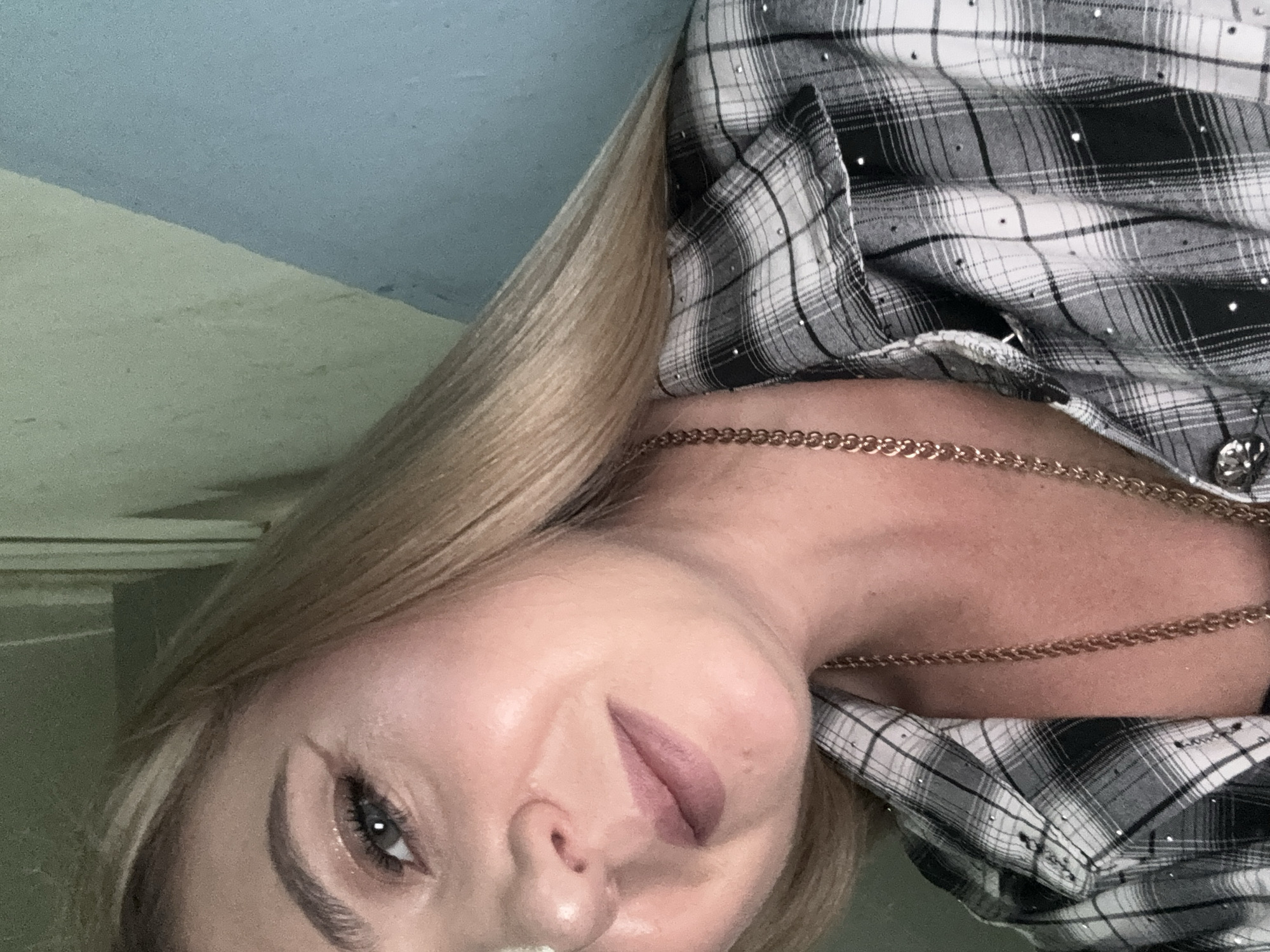 A close-up, head and shoulders, indoor, selfie-style shot features a blonde woman with a fair complexion, wearing a grey and white plaid shirt adorned with small rhinestones. She has winged eyeliner and is wearing a gold chain necklace. The background consists of a textured wall, partially painted light green and partially light blue, with a wooden beam visible in the lower left corner. The lighting is somewhat dim and diffused. There is no indication of the specific location within Makiivka, Ukraine.
