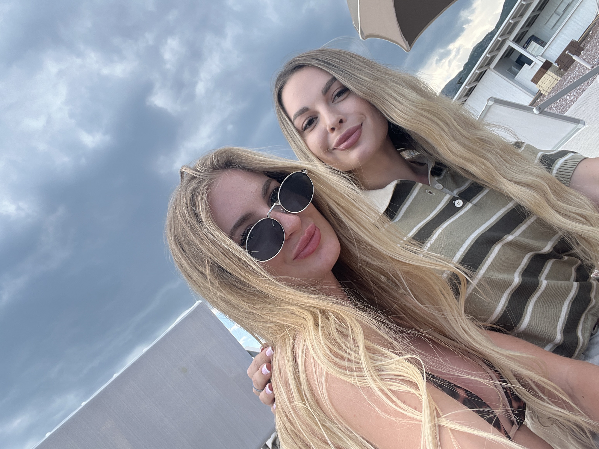 Two blonde women are posing for a selfie outdoors. The woman in the foreground is wearing sunglasses and a swimsuit, while the woman in the background is wearing a striped shirt. The background includes a cloudy sky, a building, and a beach umbrella. The location is Sveti Stefan, Montenegro.