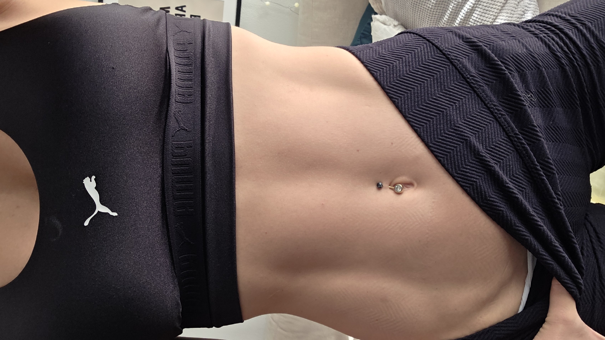The media file is a close-up, horizontal shot of a person's midsection. The individual is wearing a black sports bra with a white Puma logo on the left side and black patterned leggings. Their abdomen is toned, with visible abdominal muscles. A belly button piercing is present, featuring a circular barbell with a clear gem at the top and a dark bead below. The background is blurred, showing hints of furniture and possibly string lights, suggesting an indoor setting. The lighting appears to be soft and natural, possibly from a window. There is no specific time of day or weather information discernible. The crops highlight details of the clothing, the abdominal area, and the belly button piercing. The overall impression is one of fitness and body confidence. No legible text is visible in the main image or the crops.
