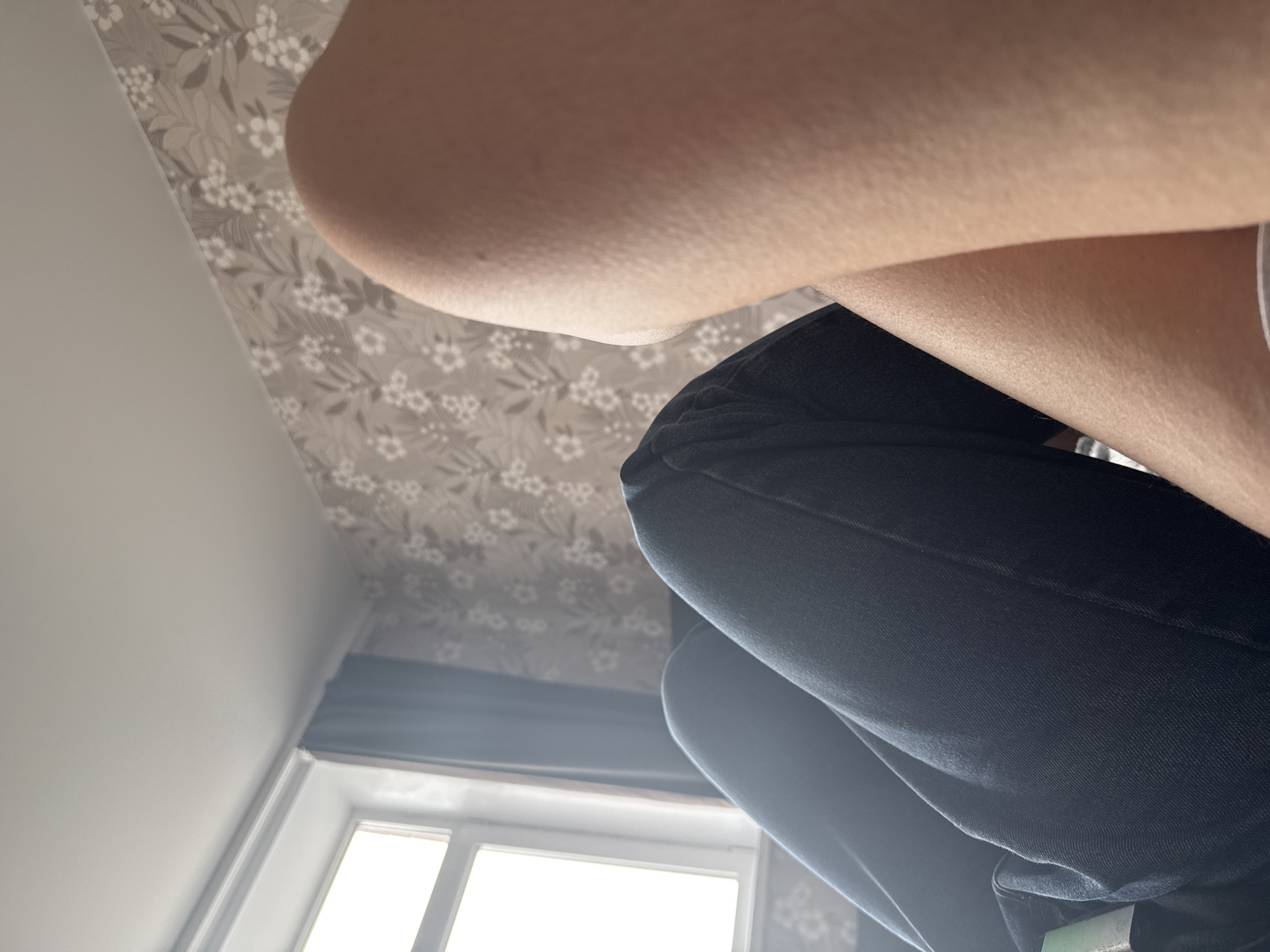 The image shows a close-up view from below, focusing on a person's crossed arms and legs clad in dark denim jeans. The background includes a white wall, a ceiling covered in a light gray floral pattern, and a window allowing natural light to enter. The location is unknown and cannot be determined from the image.