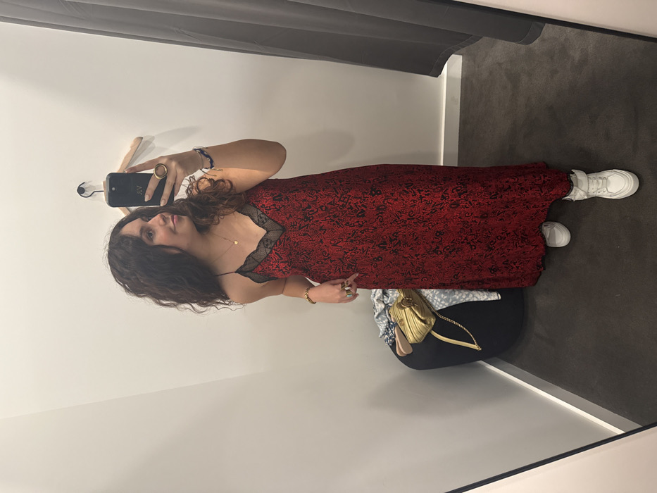A woman in a red dress with black lace trim and white sneakers stands in a dressing room. She holds a phone to take a selfie. A hanger is mounted on the wall, and a black curtain hangs from the ceiling. A handbag sits on a chair. The walls of the dressing room are white and the floor is grey.