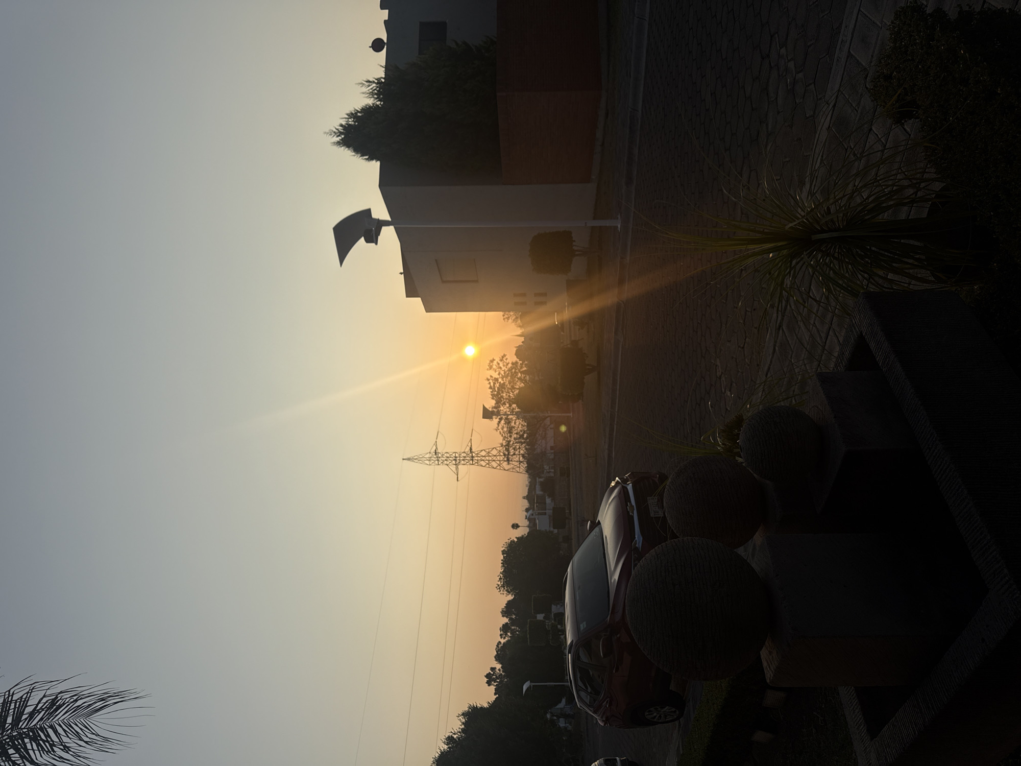 A landscape photograph shows a partially obscured sunset over a street in Heroica Puebla de Zaragoza, Mexico. Power lines and trees are visible in the background. A red car is parked on the right side of the image, partially hidden behind decorative stone spheres. The foreground is composed of a paved walkway with a succulent plant on the right. A modern building with dark brick detailing is on the left. A street light casts a soft glow.