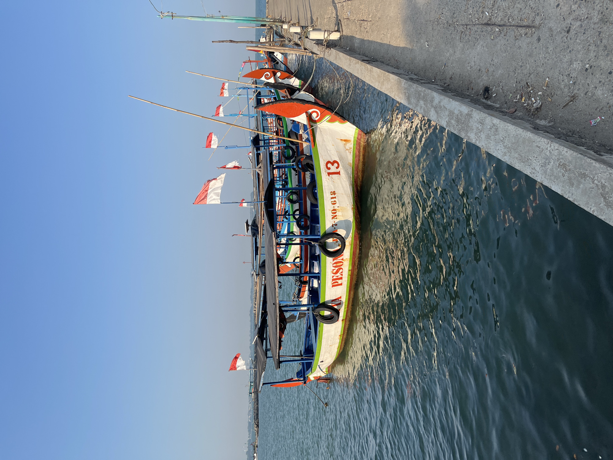 This is a photograph of a dock with several boats moored in the water. The boats are colorful and adorned with Indonesian flags. The water is calm and reflects the sky. The sky is clear and blue. The dock is made of concrete and there are some piles of debris on it. The scene is likely in Jepara, Indonesia, a city known for its maritime activities and boat building. The boats appear to be fishing boats or ferries, indicating a working harbor. The flags suggest it could be a national holiday or a patriotic display. There are no people visible in the photograph.