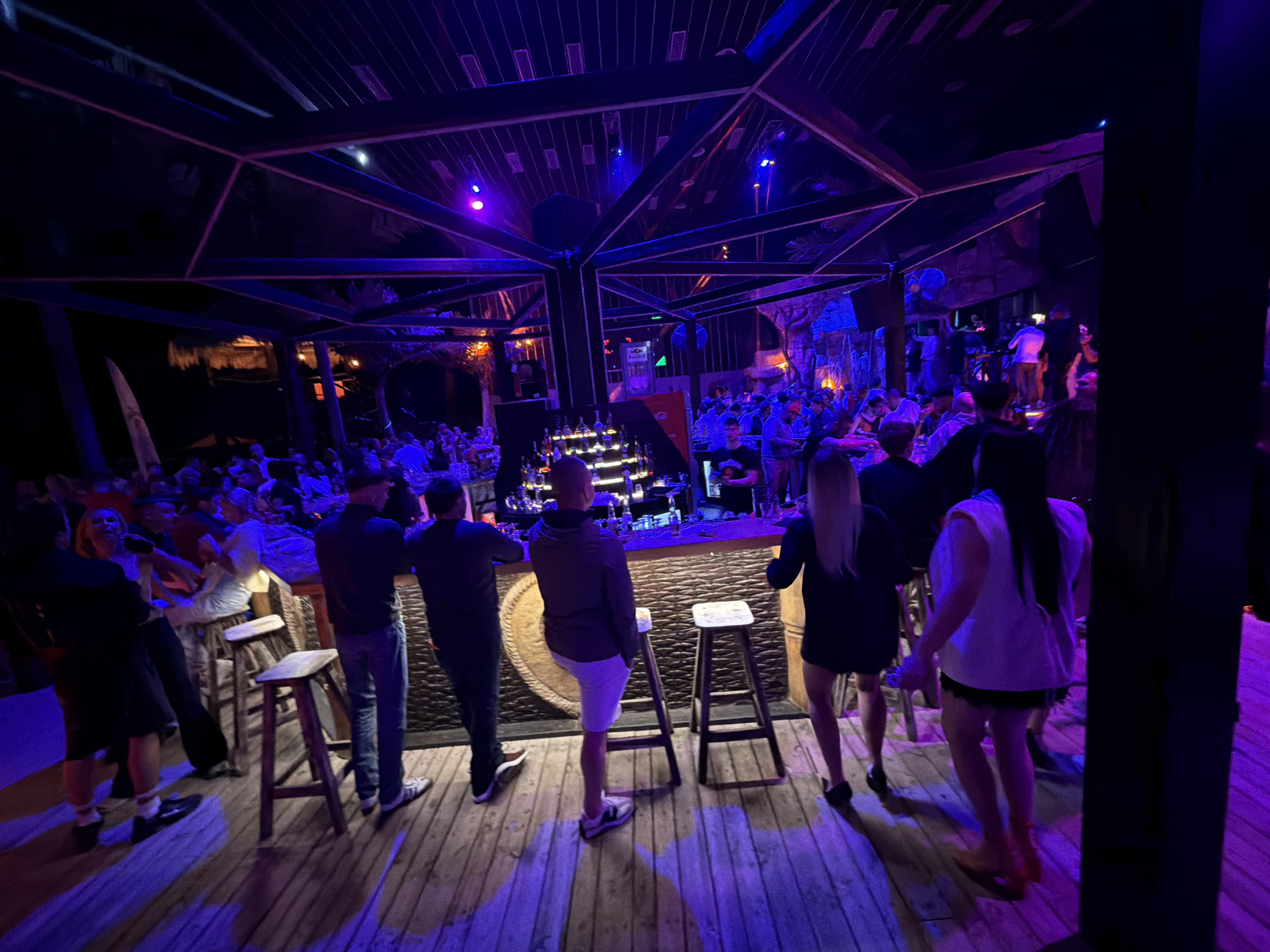 The image depicts a dimly lit, open-air bar or club, likely at night. The scene is illuminated by vibrant purple and blue lights, casting long shadows on the wooden floor. A group of people are gathered, some standing and socializing, others seated on bar stools. In the foreground, several individuals are visible from the back, facing a bar area where bottles and drinks are displayed. The architecture suggests a rustic or tropical theme, with exposed wooden beams overhead and what appears to be thatched roofing or decorative elements in the background. The overall atmosphere is lively and social, indicative of a nightlife venue. The city of Varna, Bulgaria, is not explicitly identifiable from the visual elements alone, as the scene could be from various similar locations globally.