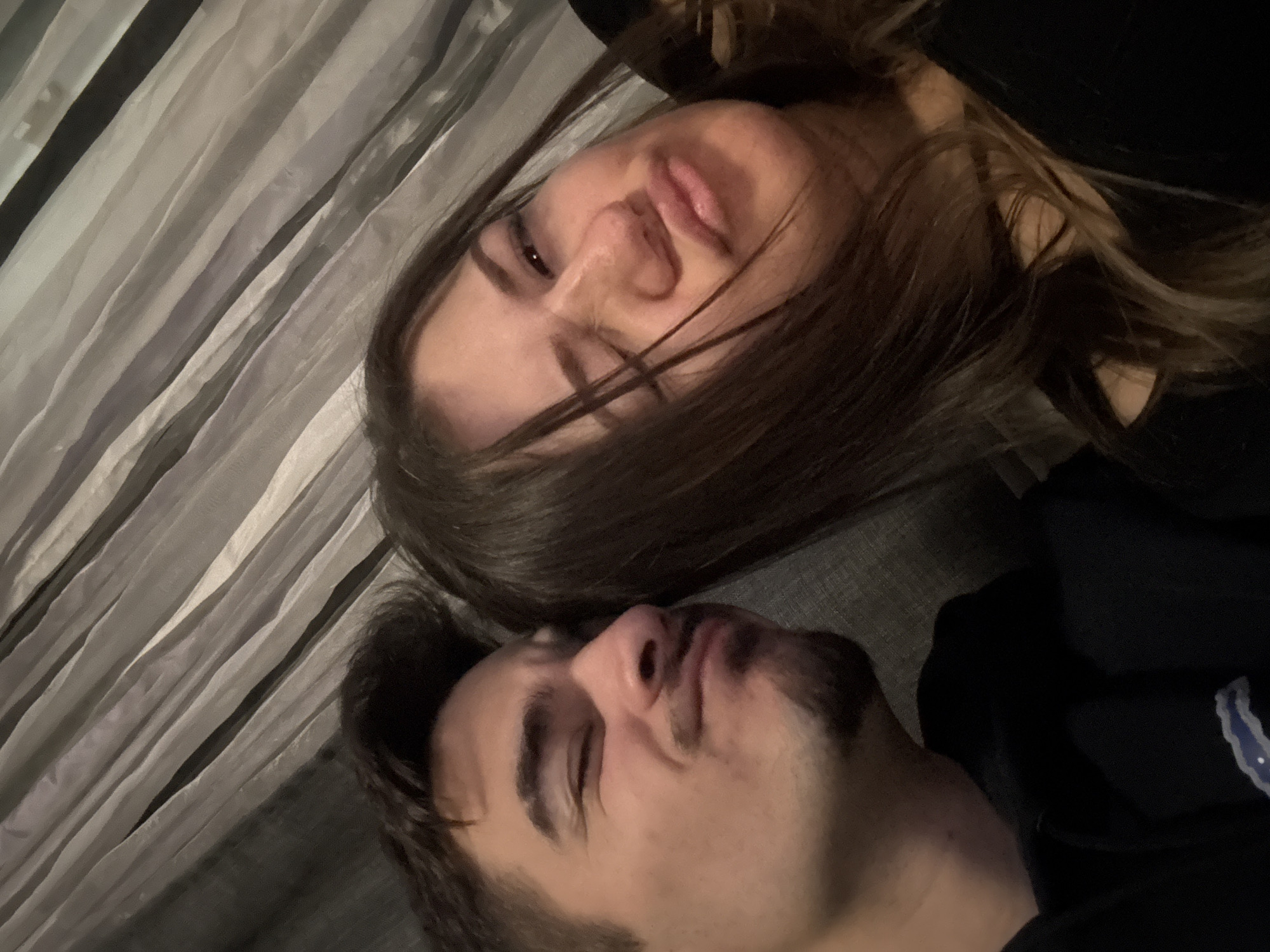 A photo of a man and a woman is shown at an unusual angle; both are lying down. The woman is at the top of the frame, and her head is tilted back, with her brown hair falling towards the camera. Her mouth is slightly open, and her eyes are partially visible. Beneath her, the man is also lying down with his head tilted, almost touching the woman's head. He has dark hair and a beard. He is wearing dark clothing. The background appears to be a striped, textured surface and dark fabric that is likely the clothing. This scene does not provide any clues to the specific location in the city of Sofia, Bulgaria.