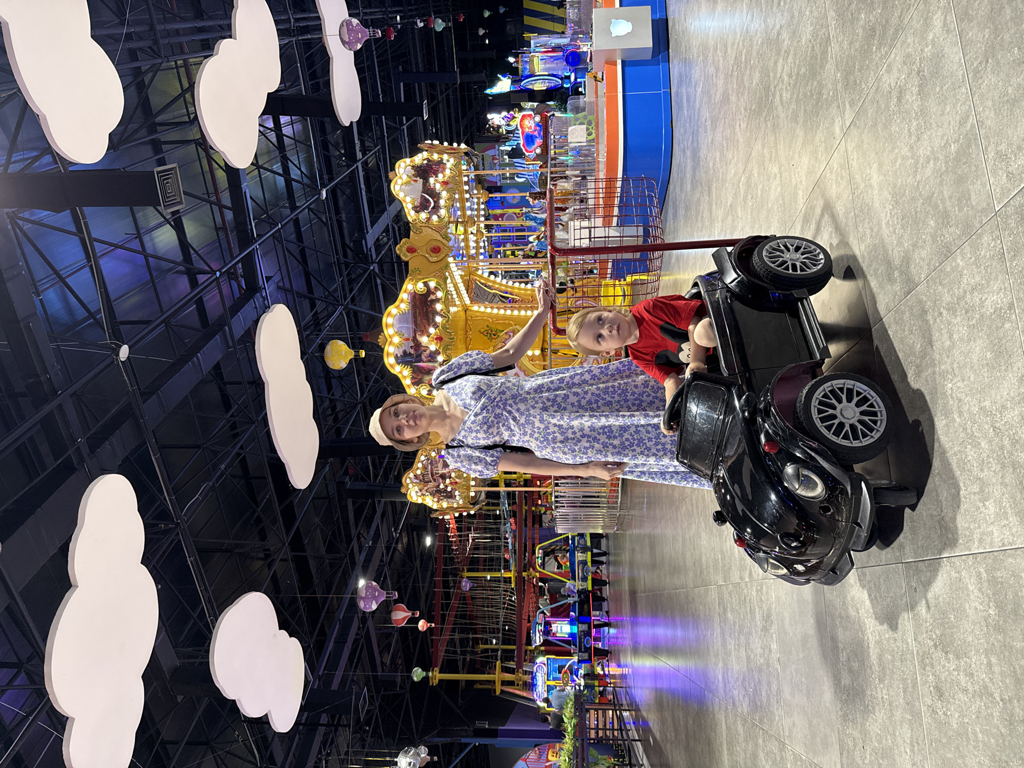 The photograph shows a woman and a young boy in a brightly lit indoor entertainment area in Tashkent, Uzbekistan. The woman, wearing a light-colored floral dress and a hat, is standing next to a black toy car. The boy, dressed in a red shirt, is seated in the car. Behind them, there is a carousel with horses, various arcade games, and an overhead ropes course. Above them, white cloud-shaped decorations hang from the ceiling. The floor is tiled in grey.