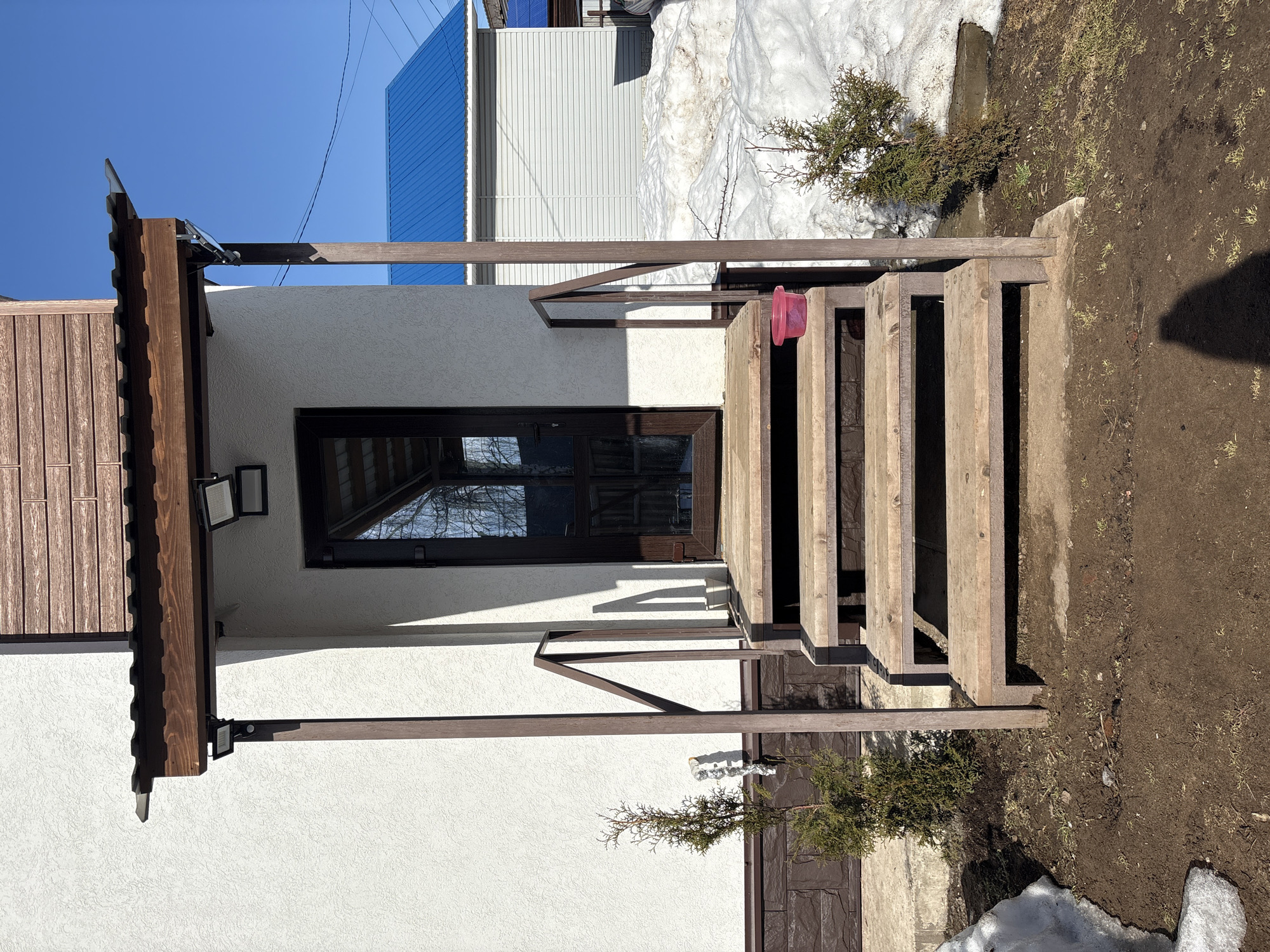 The image depicts the front entrance of a building in Votkinsk, Russia. The building features a white, textured stucco facade, with a dark brown, stone-patterned foundation extending along its base. A dark brown, glass-paneled door serves as the entrance, accessed by a set of four weathered wooden steps. Dark brown metal handrails ascend alongside the steps, supported by vertical posts of the same color. These posts also support a dark brown wooden porch canopy, topped with a dark, possibly corrugated metal roof.
Under the porch canopy, two black rectangular LED floodlights are mounted: one on the left support post, and another on the wall to the right of the door. A small white cylindrical device, possibly a security sensor or camera, is visible near the top of the left support post. On the second step from the top, a small red plastic bucket rests on the right side.
The ground directly in front of and around the steps consists of bare brown earth, showing sparse patches of emerging green grass. Patches of melting snow are present, particularly forming a larger bank to the right of the steps. Two small evergreen shrubs are planted in the ground, one to the left and one to the right of the steps. A foil-wrapped pipe extends horizontally from the wall near the left shrub. A small white pot is visible on the ground directly to the right of the steps, near the wall.
In the background, to the right of the main building, another structure with light-colored horizontal siding and a bright blue corrugated metal roof is visible, partially obscured by snow. The sky above is clear and bright blue. The scene is illuminated by direct sunlight, casting distinct shadows, including the shadow of an unseen individual in the lower right foreground. No people or active interactions are visible in the scene.
