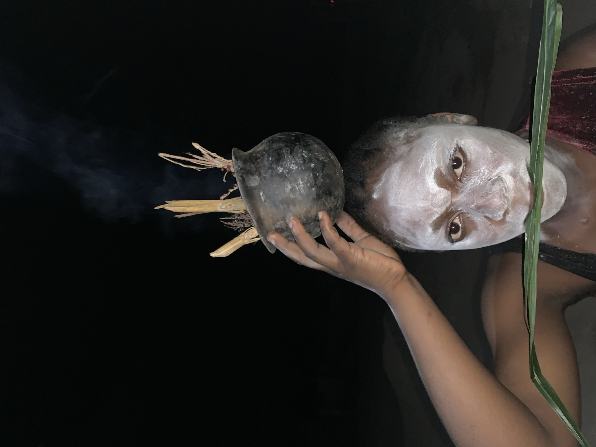 A person with a face covered in white paint is holding a dark-colored, possibly clay, pot with dried grass protruding from it. Smoke is emanating from the pot. The person is wearing dark clothing with a strip of maroon fabric visible. A long, green blade of grass is near the person's face. The background is dark. The location, Kukum, Nigeria, is not discernible from the visual information provided in the image.