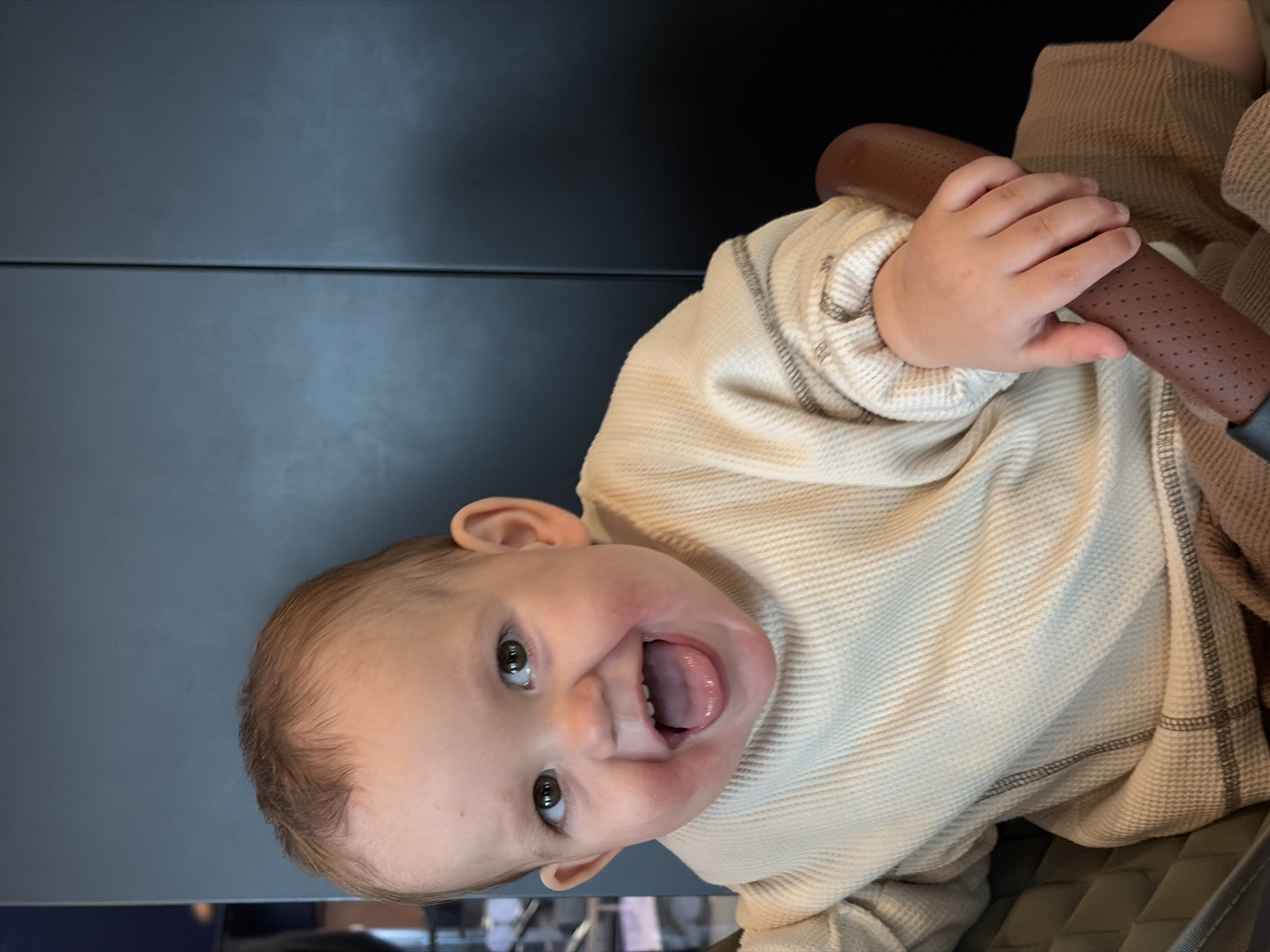 A baby, swaddled in beige waffle fabric clothing, sits in a stroller. The baby has brown hair and light skin, and is looking upwards with an open mouth. The baby's hand grips a brown, porous stroller handlebar. The stroller is set against a dark gray background that appears to be a wall. There is no indication of the location being Khimki, Russia.