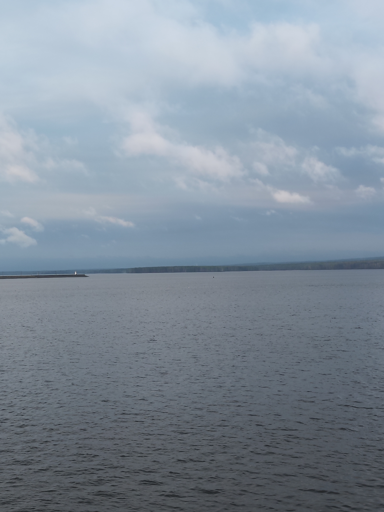 The image depicts a large body of water, likely the Kama River, under a cloudy sky. A distant strip of land is visible on the opposite shore, possibly the edge of Chaikovsky, Russia. To the left, a long pier or breakwater extends into the water, culminating in a small structure, likely a lighthouse or navigational aid. The water surface is slightly rippled, suggesting a gentle breeze.