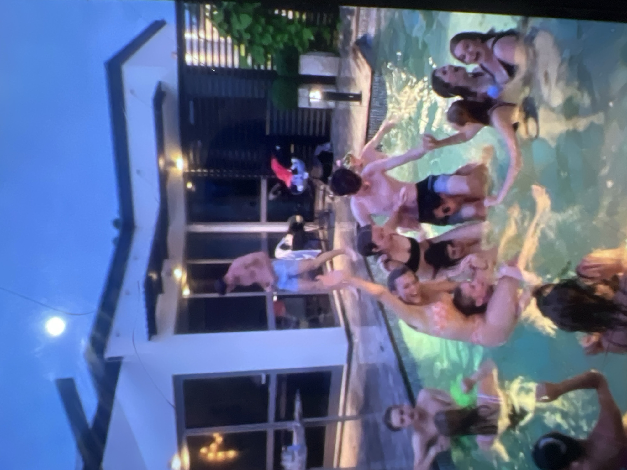 In the twilight, a group of young people are gathered in a swimming pool. The pool is illuminated by the ambient light, casting a greenish glow on the water. Several individuals are interacting, some in the water, others partially submerged. In the background, a modern house with large windows and an outdoor patio area is visible. The sky above is a deep blue, with a bright, almost full moon visible. The overall scene suggests a social gathering or party. The city of Kara-Dıykan, Kyrgyzstan, is not depicted or suggested by any elements in this image.