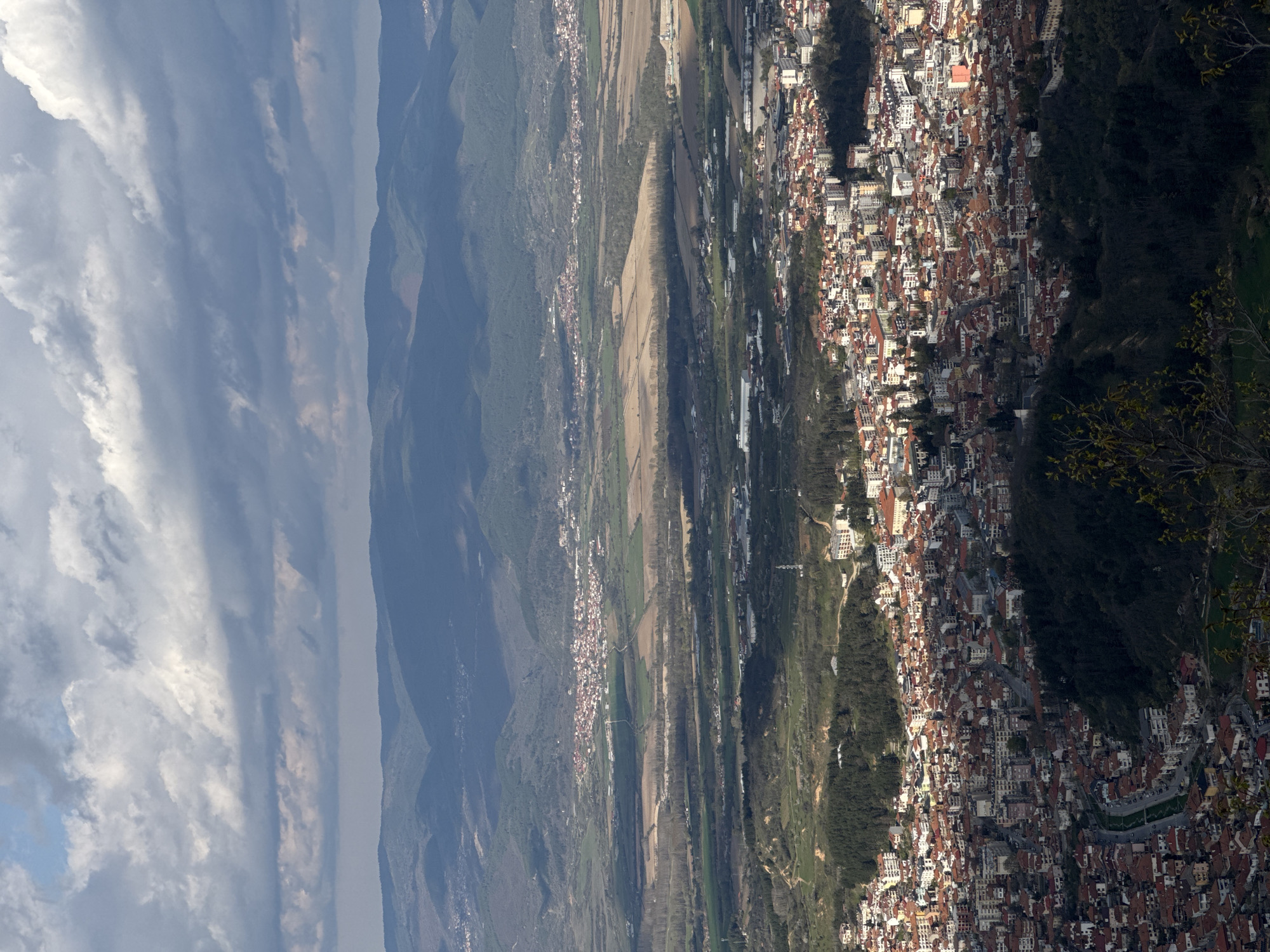 The image presents a high-angle landscape view of Delchevo, Bulgaria. The scene encompasses a valley transitioning into a dense urban area. The sky is characterized by numerous clouds. Mountains with green, tree-covered slopes are on the left and behind the city. A mix of fields and smaller settlements are visible in the valley between the mountains and the main urban area.
The city itself is a collection of tightly packed buildings, predominantly with red rooftops. Vegetation is interspersed within the urban layout, with trees and grassy areas. A winding road runs along the edge of the city near some trees on the right.