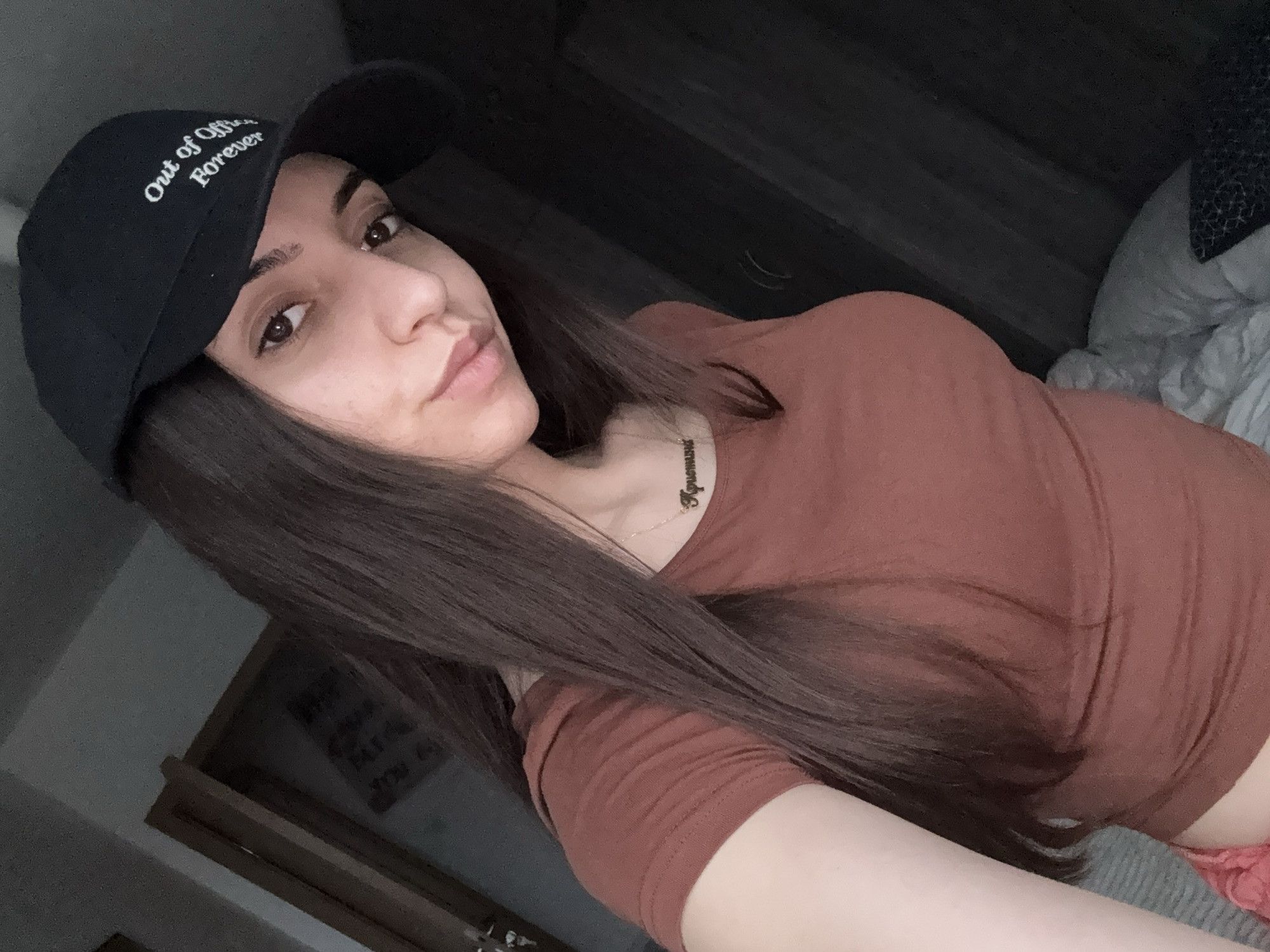 A young woman poses for a selfie-style photograph. She wears a black baseball cap embroidered with the words "Out of Office Forever," and a brown short-sleeved shirt. A delicate necklace with a nameplate is visible around her neck. Her hair is long and brown. The setting appears to be indoors, with a dark wall or wooden paneling behind her, and a glimpse of a bed with bedding to the right. A partial view of a door is seen in the lower-left corner. The location is not identifiable as Plovdiv, Bulgaria, based on the available visual information.