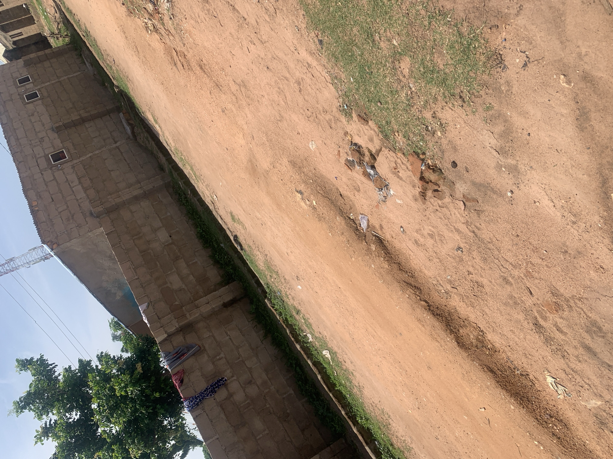 This is a photograph taken in Jauro Yinu, Nigeria. The image depicts a dirt road or pathway in the foreground, with a textured, reddish-brown surface. The road exhibits some unevenness and a slight trench or rut running diagonally across it. Patches of dry grass are visible along the edges of the road.
In the background, a brick wall constructed from concrete blocks forms a barrier. Clothing items, including a blue and white patterned garment and a red and white patterned cloth, are hung on the top of the wall. Beyond the wall, a large tree with green foliage is visible to the left. Further to the right, a building constructed of similar concrete blocks is present, featuring rectangular windows. A telecommunications tower with a lattice structure can be seen in the distance against a clear, pale blue sky. Scattered debris, including plastic and brick fragments, is present on the ground. The overall scene suggests an outdoor, semi-urban or rural environment.