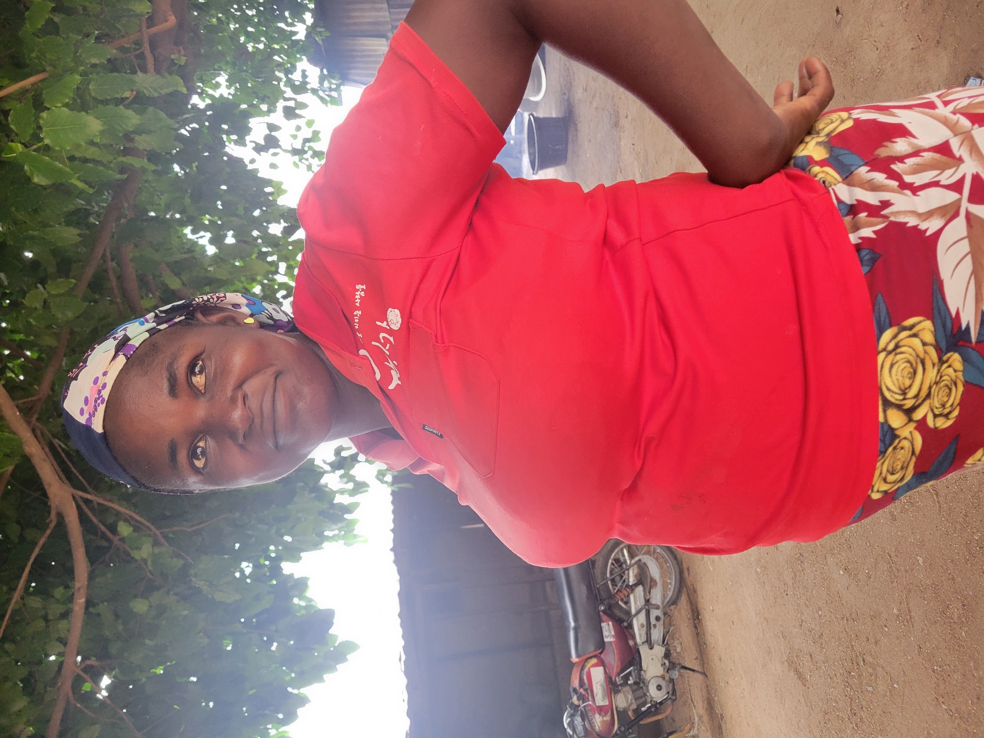 A dark-skinned woman is depicted in a full-body shot, looking directly at the camera with a confident expression and a slight smirk. Her head is tilted slightly to her right, and her hands are positioned on her hips. She is wearing a vibrant red short-sleeved t-shirt, which features white text on the chest. The text reads "MADE IN Korea" above "SS SPORTS," with larger Korean characters "사랑해요 당신" (meaning "I love you, you") and a circular emblem below. A multi-colored patterned headscarf, primarily in shades of purple, white, blue, and black, adorns her head. Her lower body is covered by a red skirt or wrap with a distinctive floral pattern of large yellow roses and green and blue leaves.
The setting appears to be outdoors in a sunny daytime environment. Lush green trees and foliage are visible in the upper left and background, indicating a natural surrounding. The ground beneath her is bare dirt. In the mid-ground to her right, there is a simple dark-roofed structure, and parked nearby is a red and white motorcycle or moped. A black bucket is also visible on the ground. The bright overhead lighting suggests it is mid-day. The overall scene, including the natural elements, simple structures, and dirt ground, is consistent with an outdoor setting in Madalla, Nigeria.