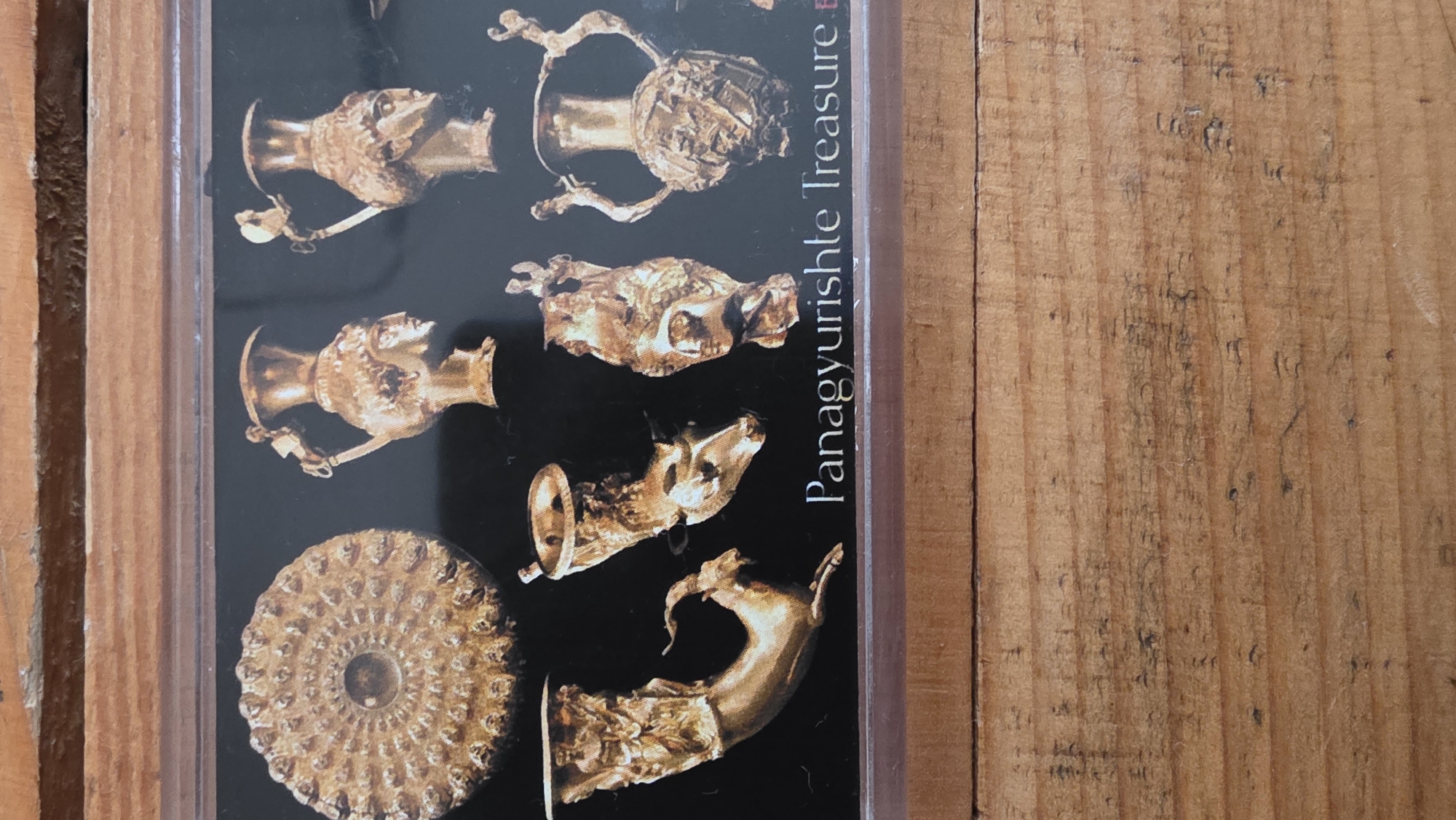 A rectangular image, likely a postcard or framed photograph, displays a collection of ancient golden artifacts against a dark background. The artifacts include several ornate rhytons (drinking vessels) shaped as animal heads, such as deer and goats, and human female busts, along with a large, intricately ribbed golden phiale or dish with a central boss. These items are characteristic of the Panagyurishte Treasure.
Running vertically along the right edge of the artifact display, the text "Panagyurishte Treasure" is clearly visible, followed by a partially obscured letter. The display is placed on a light-brown wooden surface, possibly a table or floorboards, featuring prominent wood grain, knots, and darker streaks, suggesting a rustic indoor setting. The lighting highlights the metallic sheen of the gold and the texture of the wood. No people or activities are present. The artifacts appear well-preserved, showcasing their detailed craftsmanship. The location context of Blagoevgrad, Bulgaria, aligns with the Panagyurishte Treasure, which is a significant archaeological find from Bulgaria.