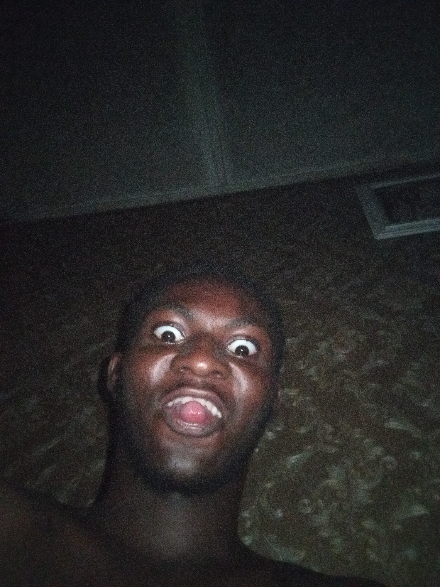 A close-up, low-angle photograph depicts a dark-skinned individual making an exaggerated facial expression, eyes wide and mouth open. The background consists of patterned wallpaper and a ceiling with what appears to be a recessed light fixture. The location is unknown.
