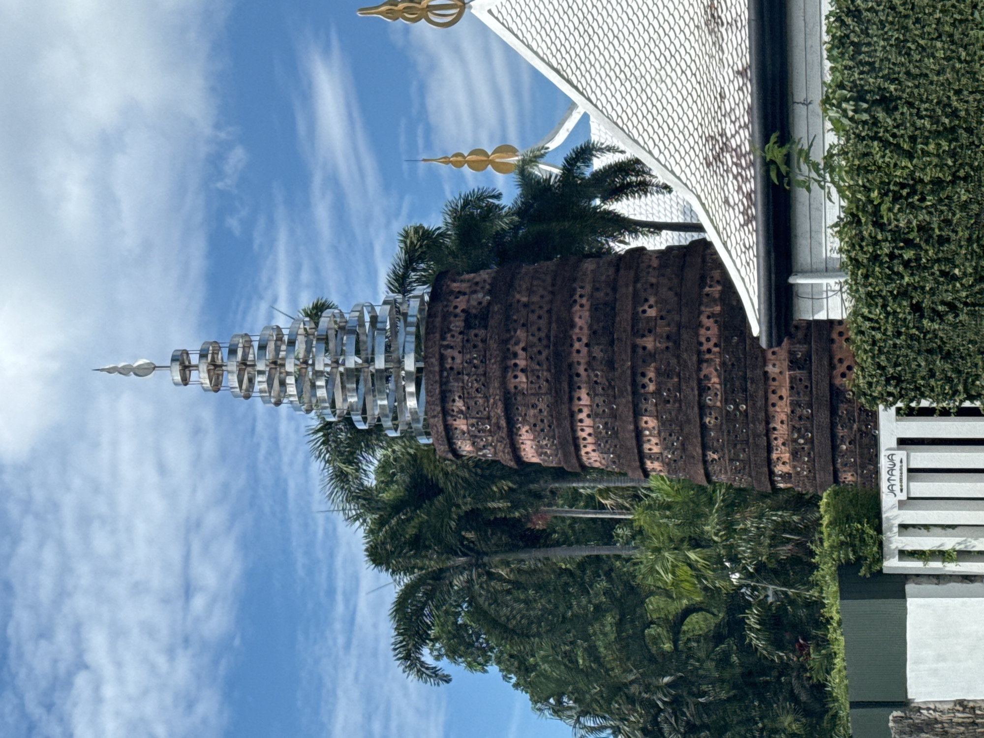 A brick tower with a metal spire stands in front of palm trees and a white-tiled roof. The tower is dark brown and appears to be made of many stacked rings. The metal spire is silver and intricately shaped. The roof has a distinctive curved shape, and there are golden finials on its peaks. The sky is blue with scattered clouds. In the foreground, a white fence with a sign that reads "JATAWA" is visible, along with lush green bushes and a stone wall.