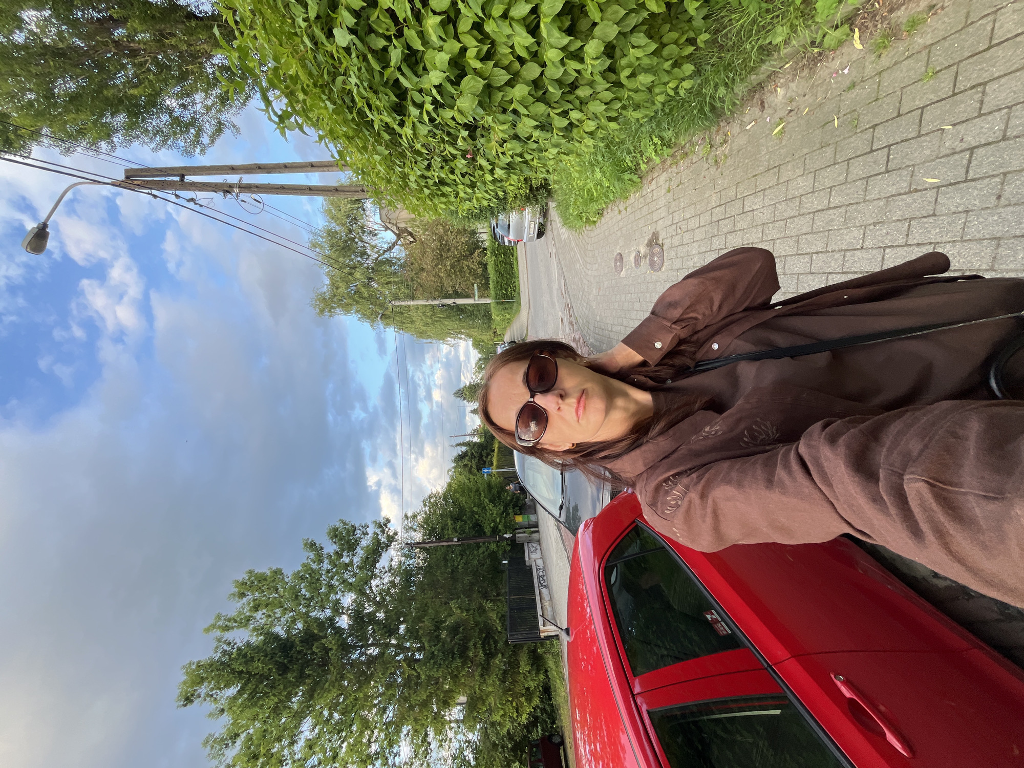 The image captures an outdoor selfie of an adult female on a paved sidewalk, likely in a residential area. The woman is in the foreground, angled slightly from below, wearing dark oval sunglasses and a long-sleeved brown shirt or jacket with subtle embroidered details on the upper left arm. Her brown hair is parted on her left, and she holds the camera with her left arm extended while her right hand is positioned at the back of her neck or hair.
To her immediate left, the rear and side of a bright red hatchback car are visible, parked parallel to the sidewalk. A white "Z" decal is present on the car's rear window. Behind the red car, a dark-colored sedan or hatchback is also parked. Further to the left, behind the parked cars, a black metal gate and fence are discernible, backed by dense green trees and foliage. Unidentifiable graffiti is visible on a low wall or curb behind the red car.
The sidewalk, paved with rectangular grey pavers, extends into the mid-ground, curving gently to the right. Three circular metal utility covers are embedded in the sidewalk near the woman's feet. Along the right side of the sidewalk, a tall, thick hedge of vibrant green leaves forms a continuous barrier. Beyond the hedge, a narrow strip of grass and weeds runs along the sidewalk's edge.
In the mid-ground on the right, a tall wooden utility pole stands with multiple electrical wires and insulators. A white street lamp with a curved arm is attached to the top of this pole. Another, slightly obscured wooden pole is visible behind the first. Further down the street, a silver car is parked facing away from the viewer. The background is characterized by numerous green trees and foliage, including a large deciduous tree on the far left and a weeping willow-like tree further down the street on the right. The sky overhead is partially cloudy, with areas of light grey clouds and patches of blue sky, particularly towards the upper right, where a small dark speck, possibly a bird, is visible. The overall lighting appears diffused, suggesting an overcast day or late afternoon/early evening. The location is a typical Unknown residential street.