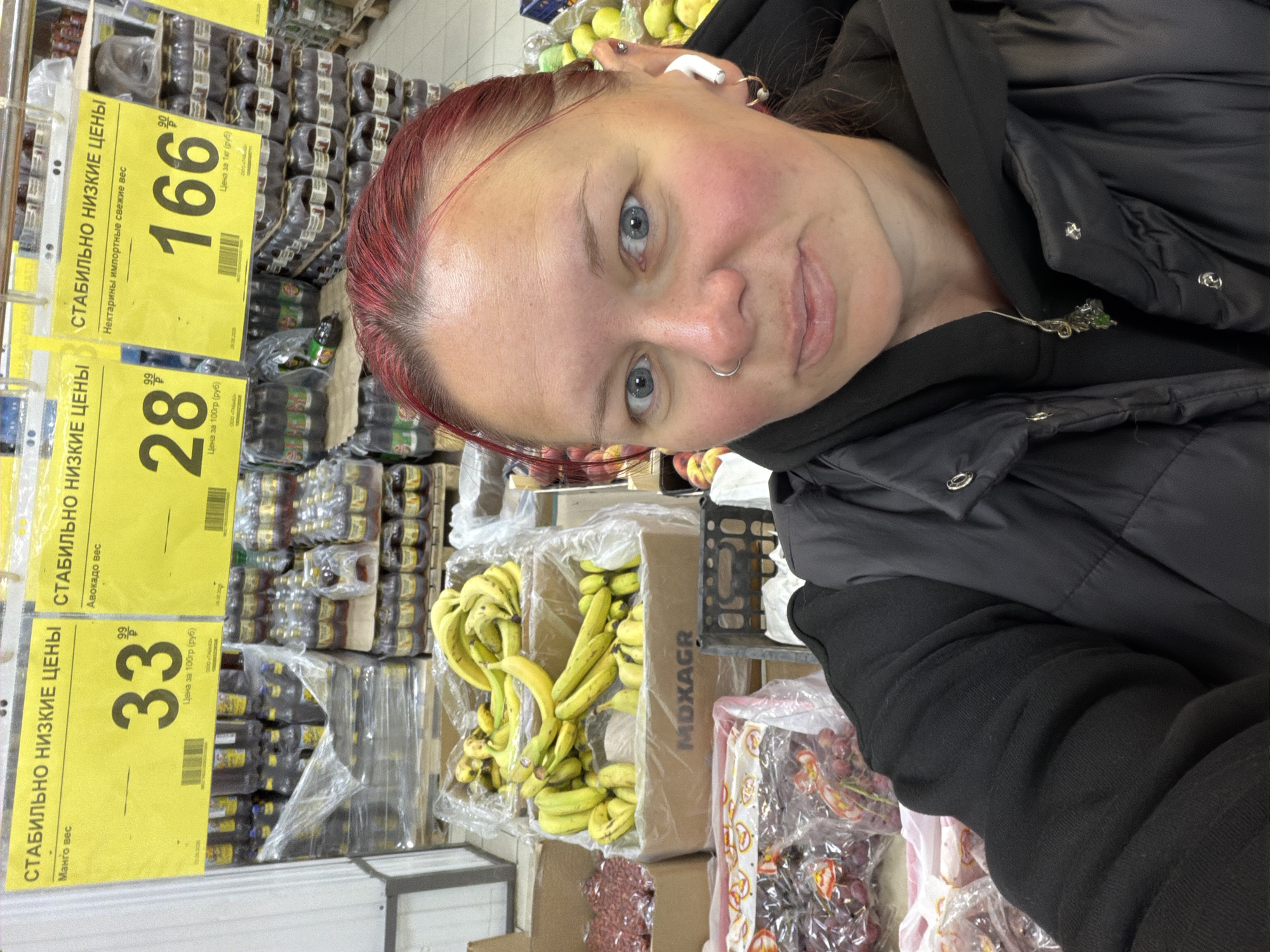 A woman takes a selfie at a grocery store in Chaikovsky, Russia. The woman is fair-skinned with blue eyes and dyed red hair. She is wearing a black jacket and a silver nose ring. She also has an earbud in her left ear. In the background are shelves of groceries, including bananas, grapes, mango, and avocado. There are also yellow price tags with Cyrillic text, showing prices in rubles.