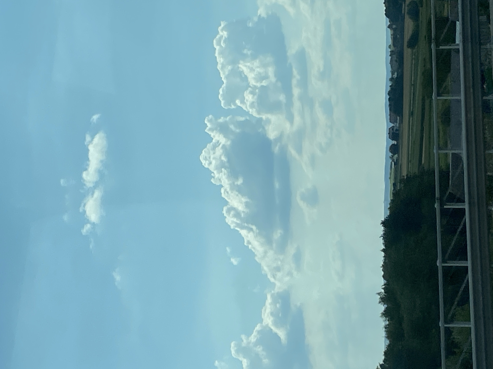 The image depicts a panoramic view of a rural landscape under a bright, partially cloudy sky. In the foreground, a light-colored railing runs horizontally across the bottom of the frame, suggesting the perspective is from a bridge or elevated walkway. Beyond the railing, a dense line of dark green trees forms a backdrop, leading the eye to a rolling landscape of green fields, some with patches of brown, indicating agricultural activity. Several buildings, including houses with dark roofs, are scattered across the mid-ground, with more distant hills fading into the horizon. The sky is a vibrant blue with scattered white, fluffy cumulus clouds. The overall impression is one of a serene, sunlit countryside, possibly in or around Zamiechów, Poland, given the characteristic architecture and landscape often found in Polish rural areas. There are no people or animals visible in the image, and no action or interaction is depicted.